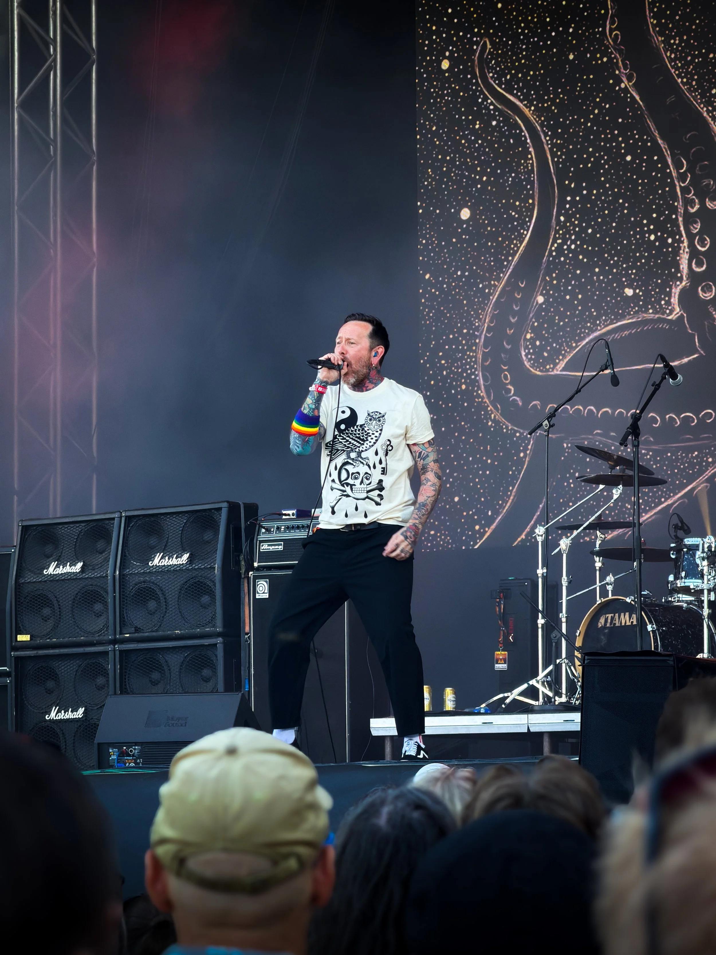 A man with tattoos on his arms and neck, speaking into a microphone on a stage during a live music performance. He is wearing a white T-shirt with black graphics and black pants, and has colorful rainbow wristbands on his right arm. The stage features multiple Marshall speakers and musical equipment, with a large screen behind displaying abstract, dotted art in warm tones.