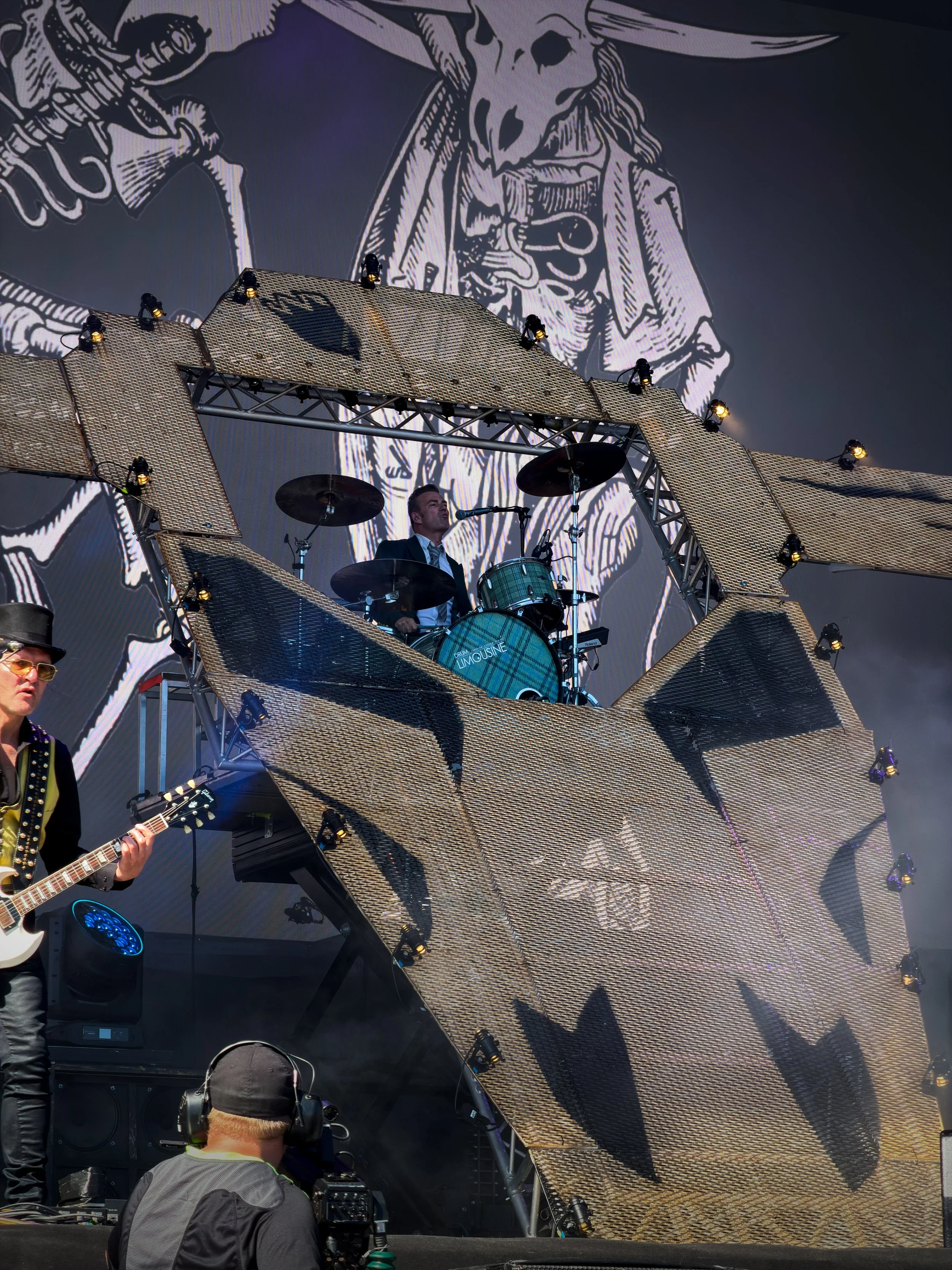 Live music performance with a drummer and a guitarist on stage, featuring a large digital backdrop with a stylized drawing of a woman with a skull face. Stage setup includes metallic scaffolding and lighting.