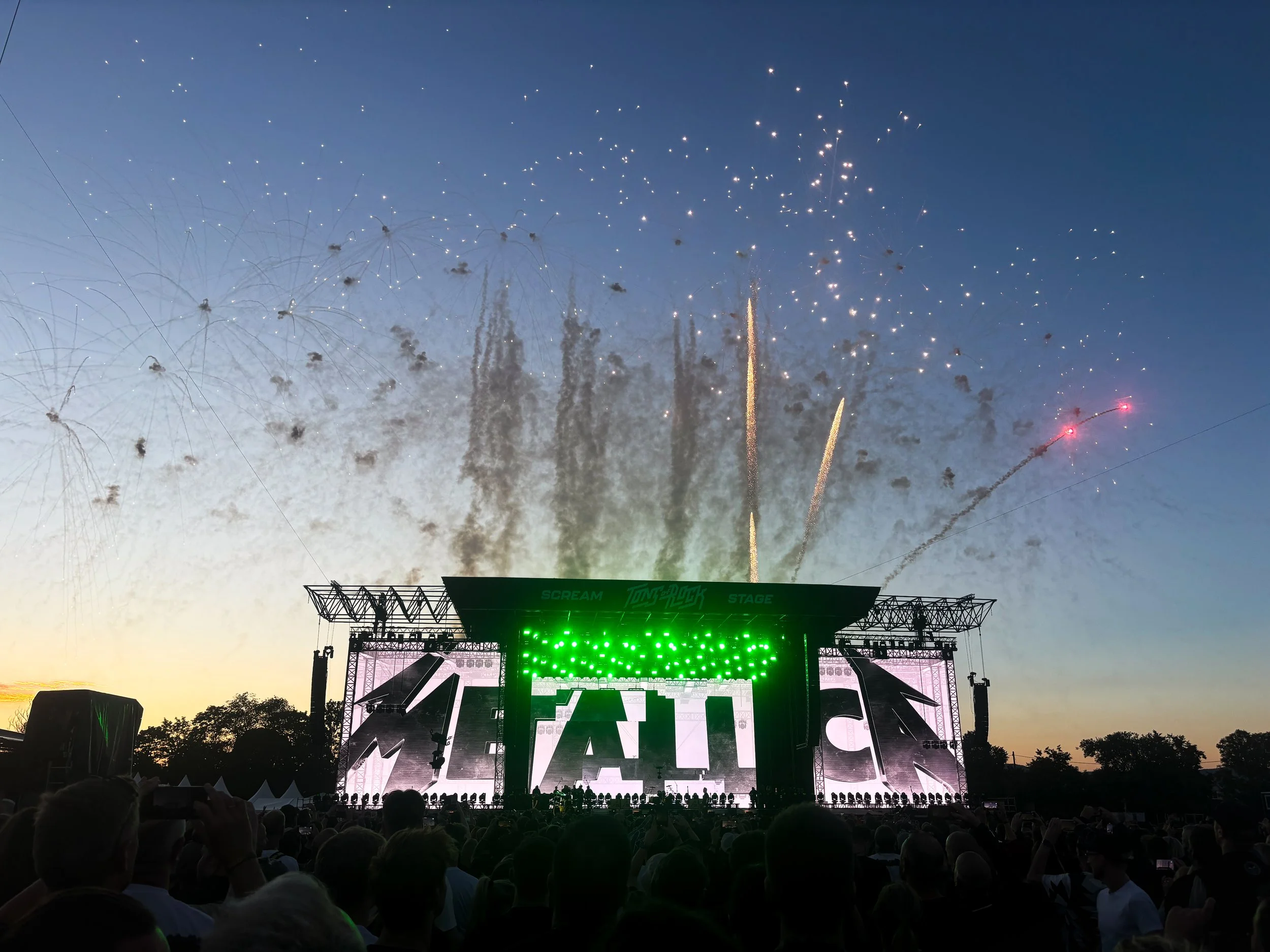 Crowd watching a concert on an outdoor stage with fireworks in the sky at sunset.