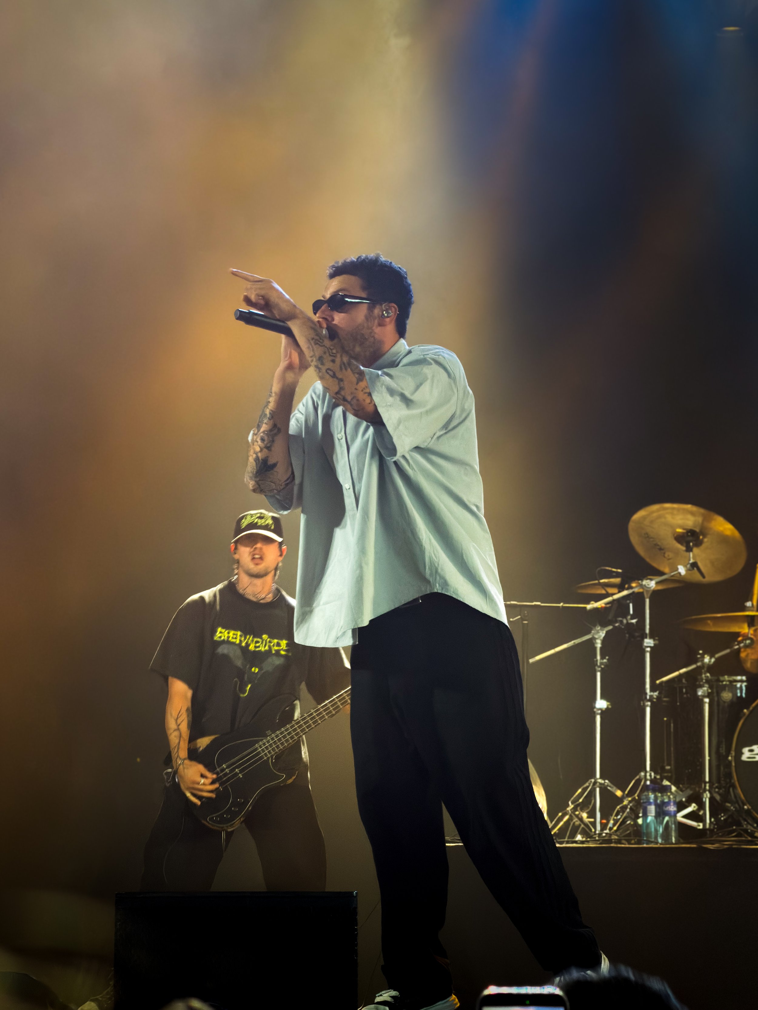 A male performer wearing sunglasses and a loose white shirt, singing into a microphone on stage with arms raised, and tattoos visible on his arms. Behind him, another person is playing an electric guitar, and musical equipment including drums and cymbals are seen in the background. Stage lighting creates a hazy atmosphere.