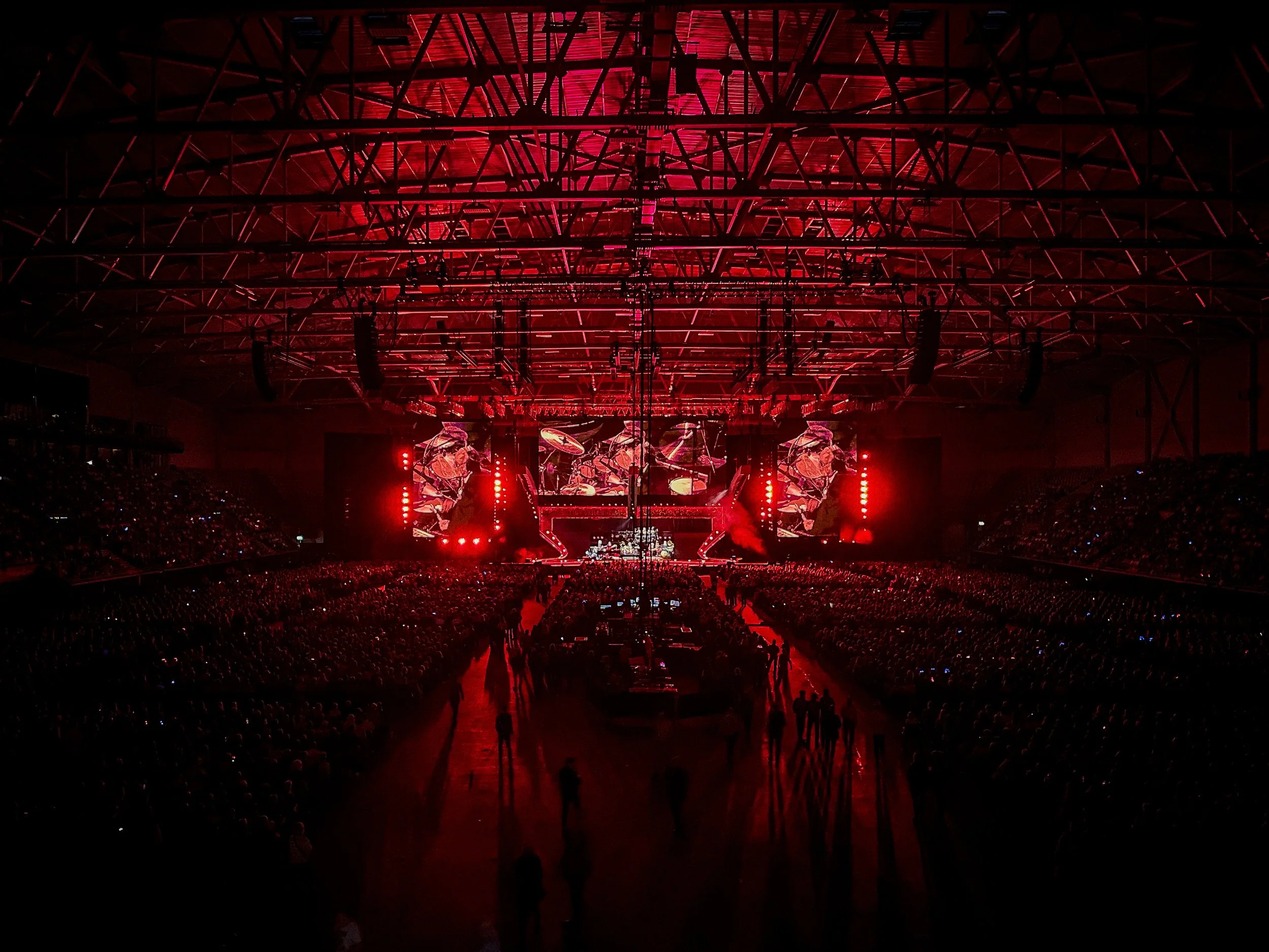Concert venue with a large crowd, stage, and red lighting illuminating the ceiling's metal framework.