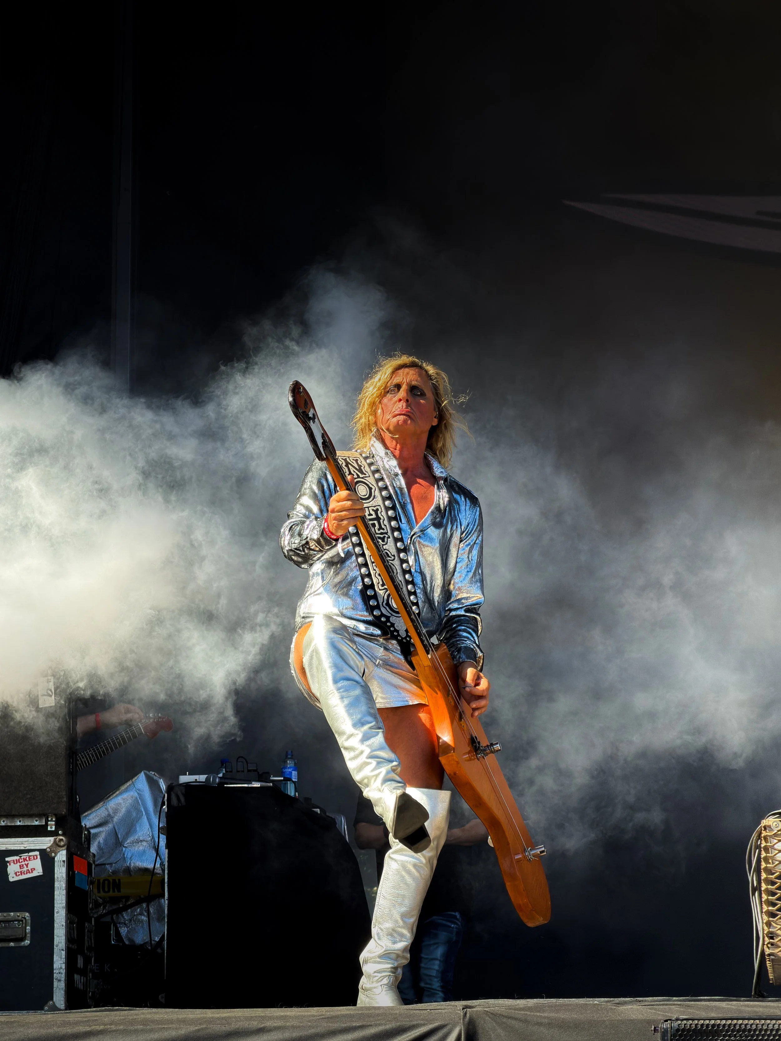 A performer dressed in a shiny silver outfit on stage, holding an orange electric guitar, with a backdrop of stage smoke and dark background.