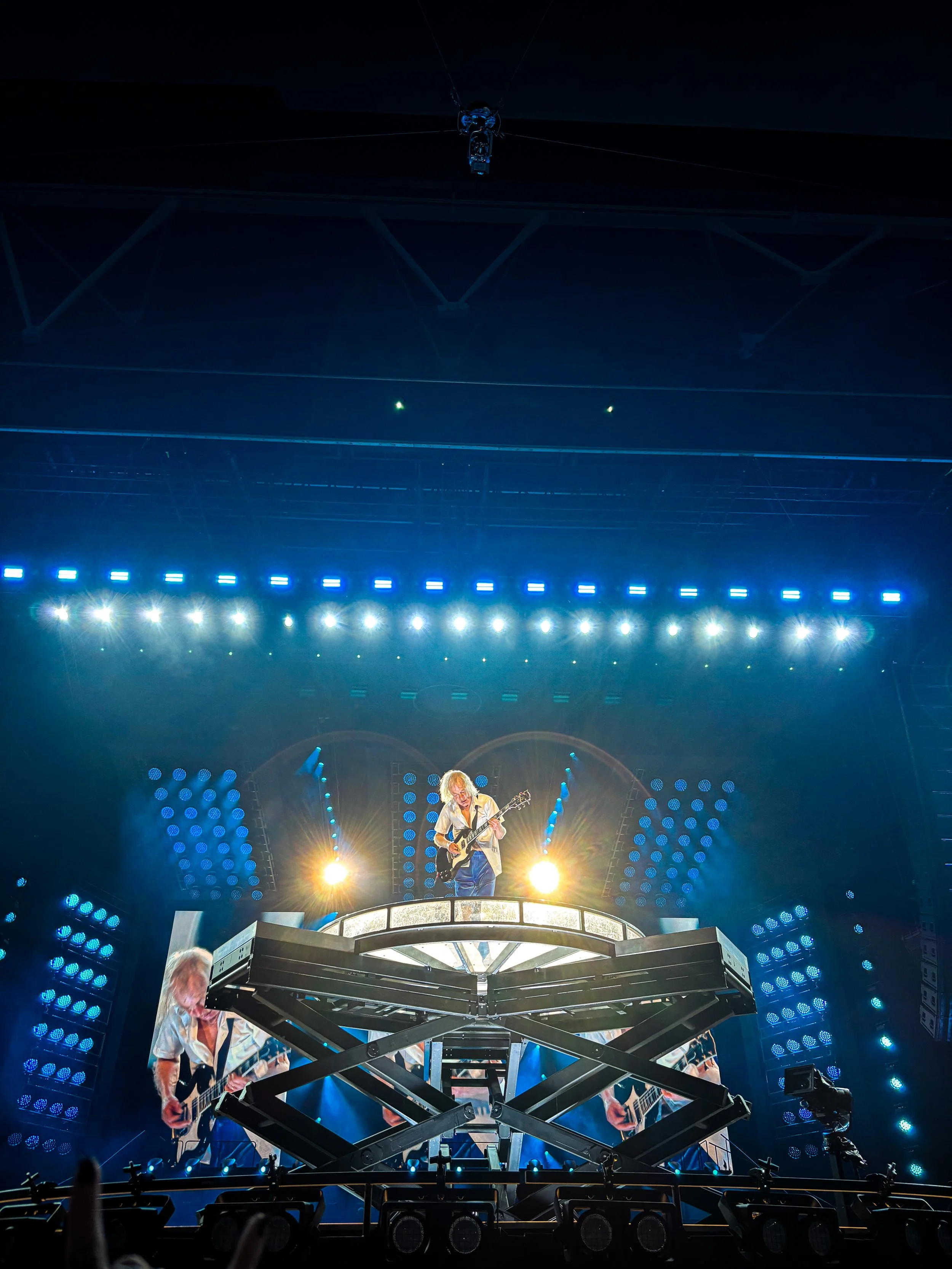 Musical performance on a brightly lit stage with musicians playing guitars and keyboard, large screens displaying the performers, and audience members in view.