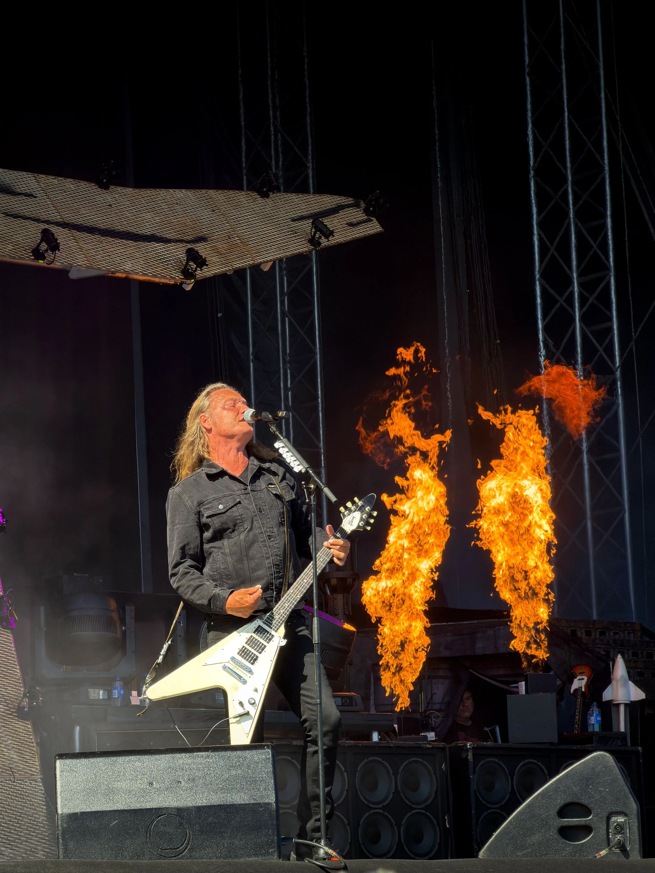 A musician with long hair playing an electric guitar on stage with flames rising in the background during a performance.