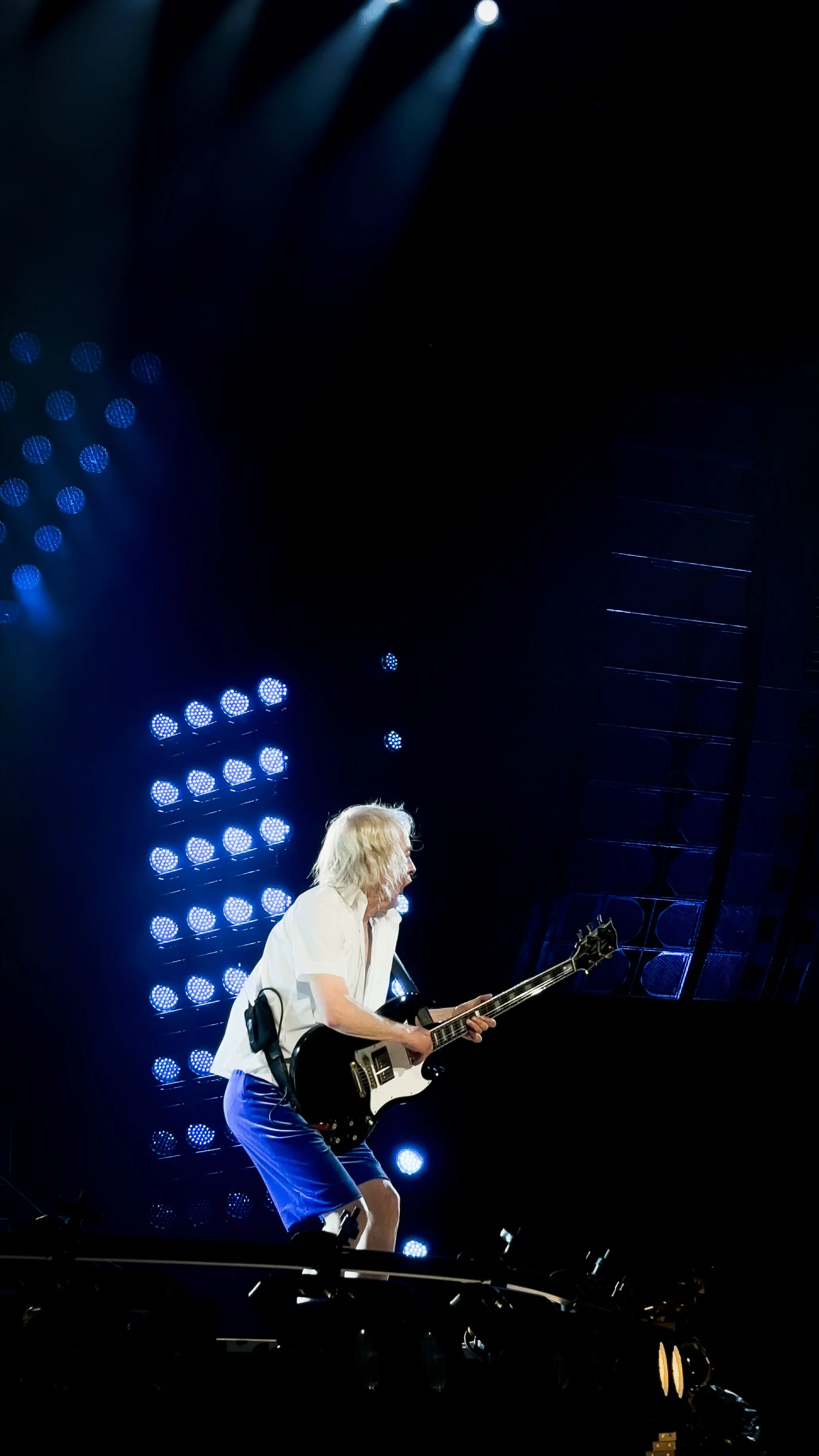 A man with long white hair playing an electric guitar on stage, lit by numerous blue spotlights.