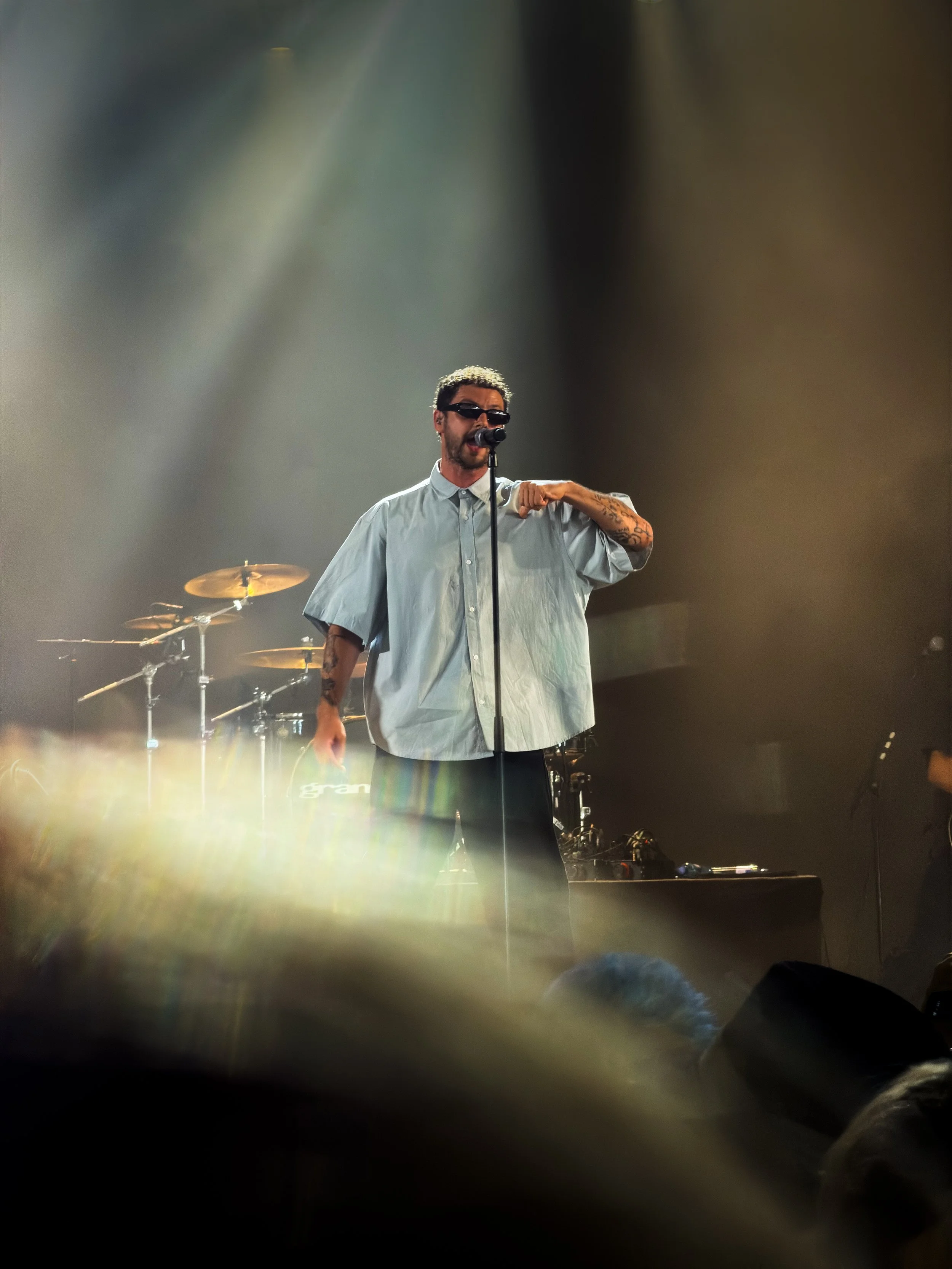 A male performer with tattoos and sunglasses singing into a microphone on stage, with a drum set in the background, under stage lighting.