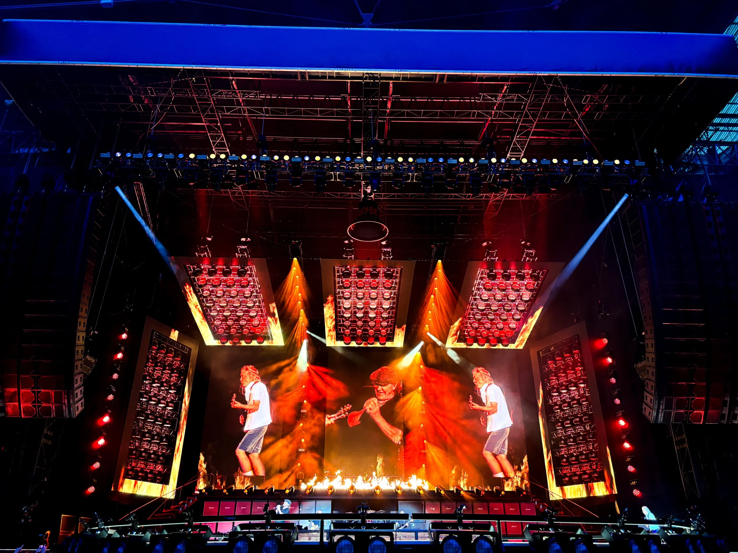 A stage with bright lights, large screens displaying musicians, and people performing during a concert with flames and colorful lighting.
