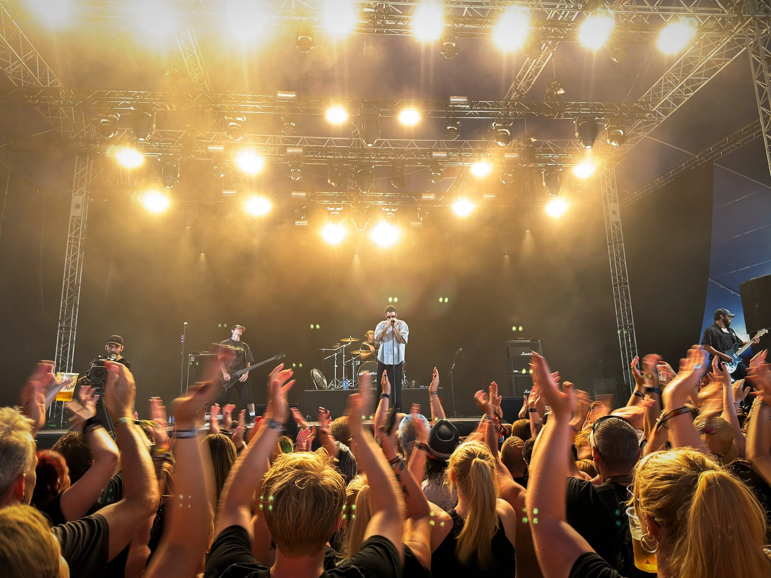 A live concert with a band performing on stage under bright yellow stage lights. The audience is cheering and raising their hands, some holding smartphones and cameras. The band includes at least four members: a singer, guitarists, and a drummer. The stage is set with large speakers and musical equipment.