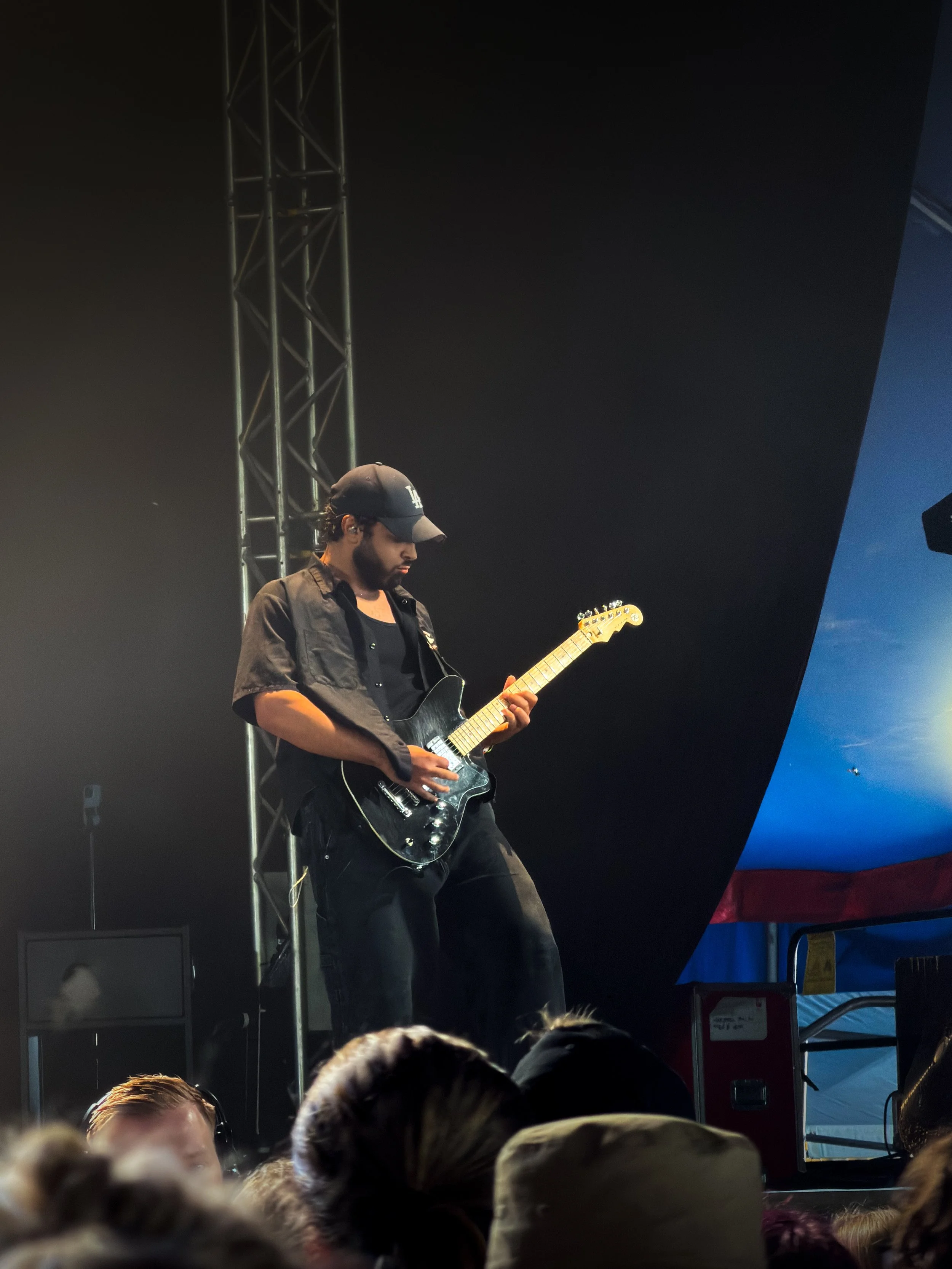 A musician wearing a black cap and black jacket playing an electric guitar on stage during a concert, with a metal support structure and a dark backdrop.
