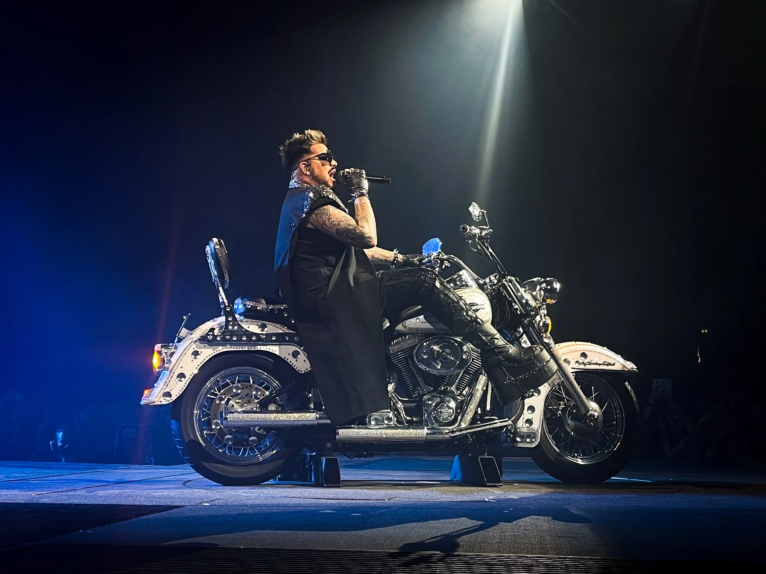 A man withstyled hair, sunglasses, tattoos, and gloves, sitting on a shiny, chrome motorcycle on a stage with dark background and spotlight.