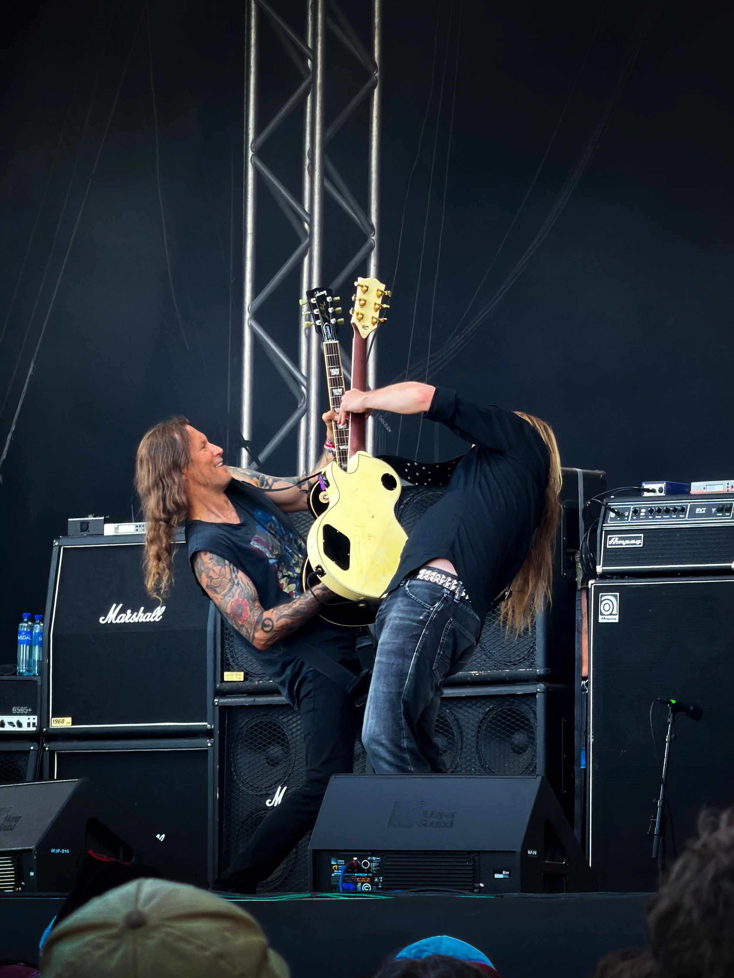Two musicians performing on stage, one playing an acoustic guitar and the other leaning over, with various amplifiers and equipment in the background.