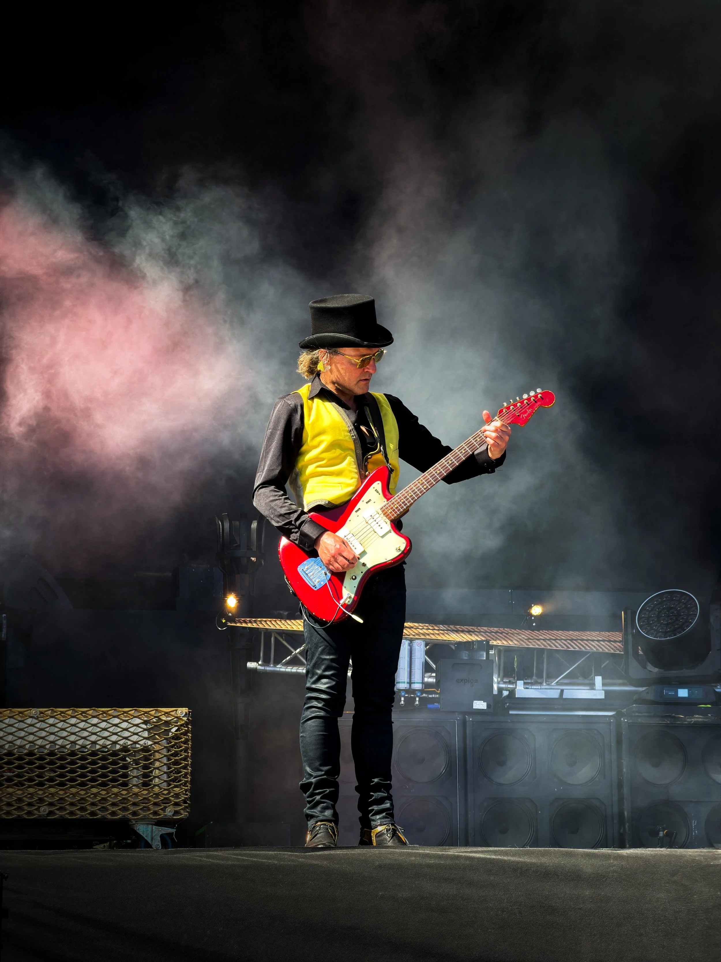 A man wearing a black top hat and sunglasses playing a red electric guitar on stage with fog and stage equipment in the background.