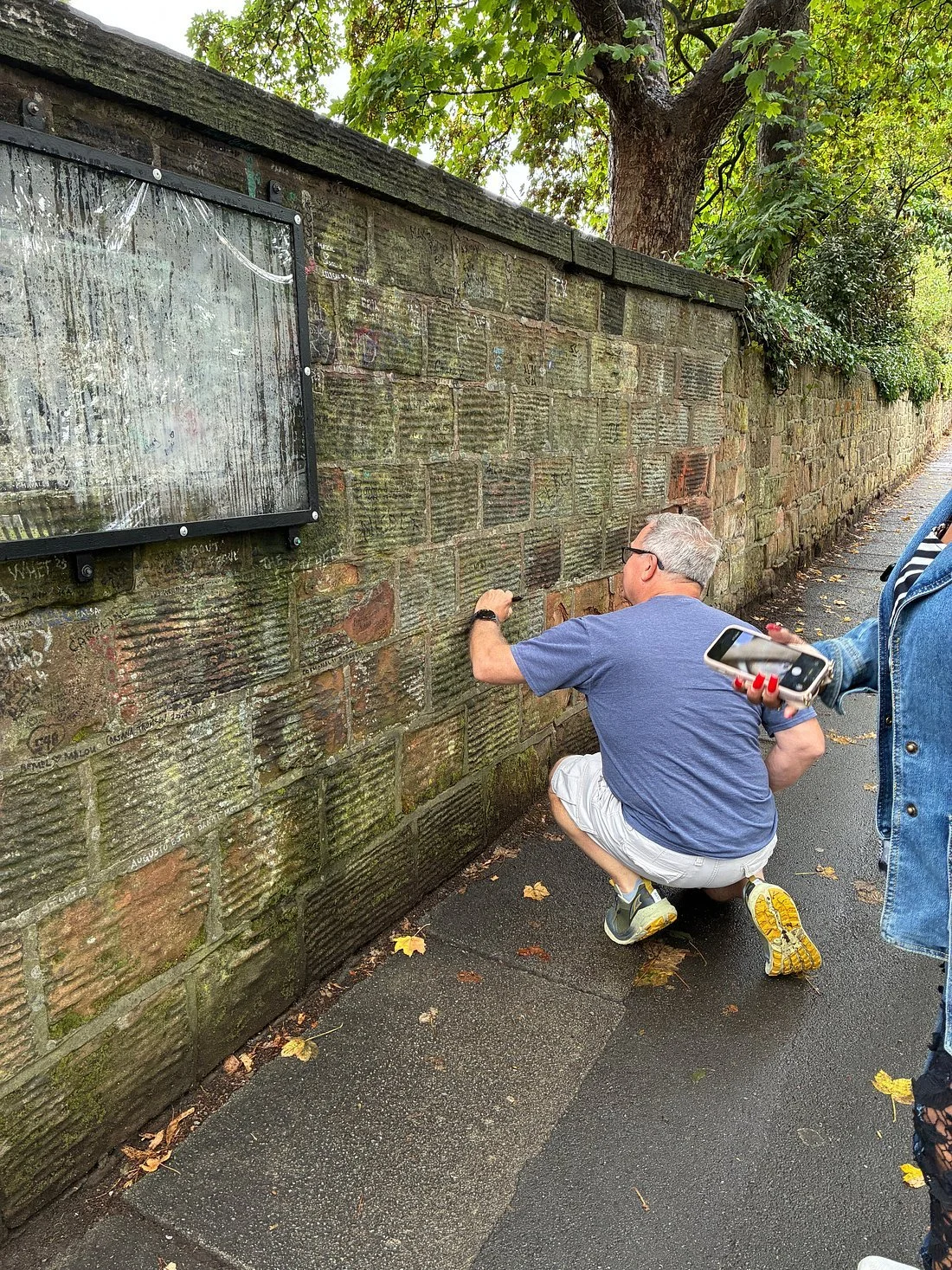 Man reading plaques on mossy brick wall while another person holds smartphone on Penny Lane Liverpool