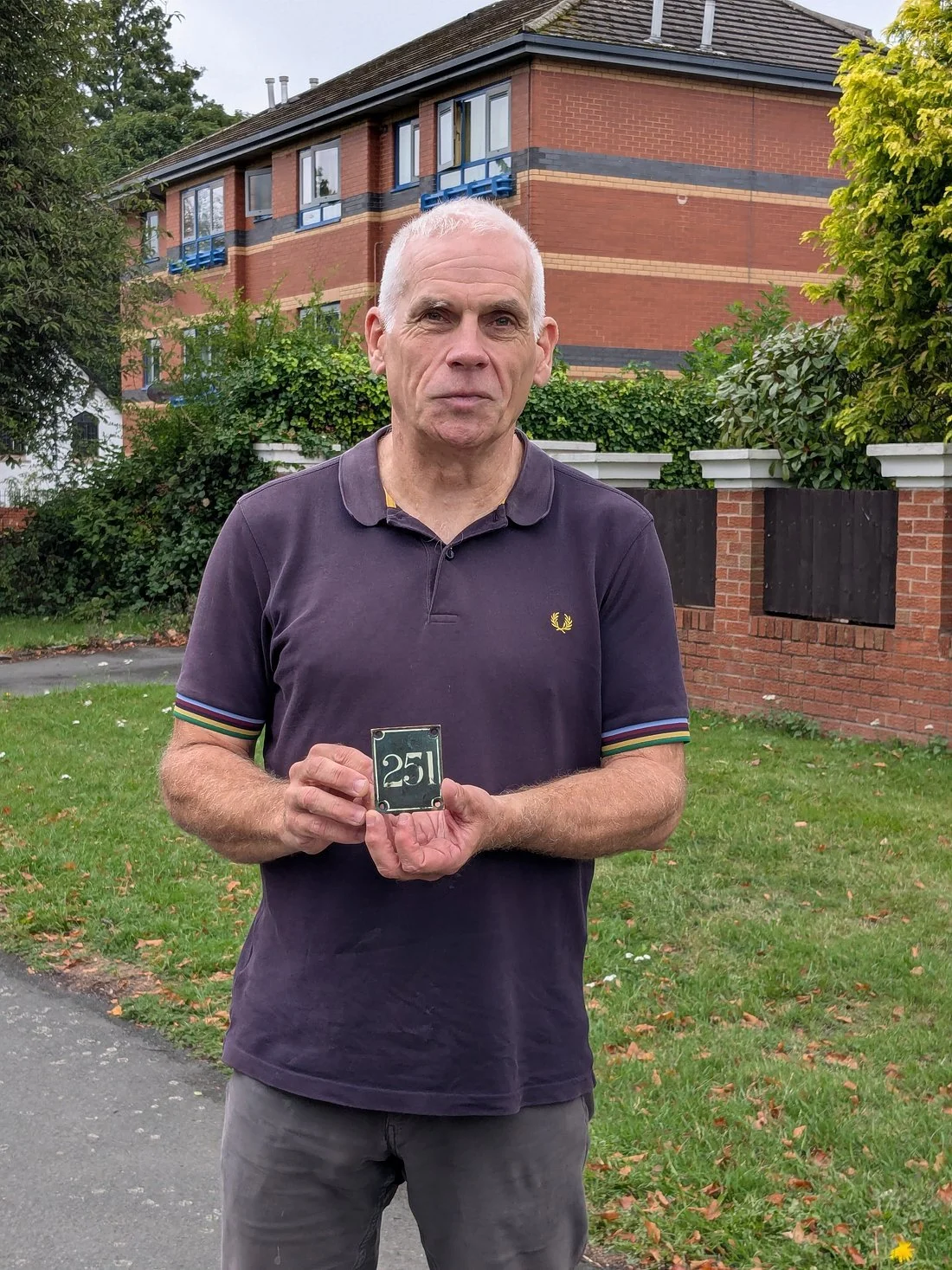 Tim standing on a grassy area in front of a Liverpool brick house, holding a small plaque with the number 251