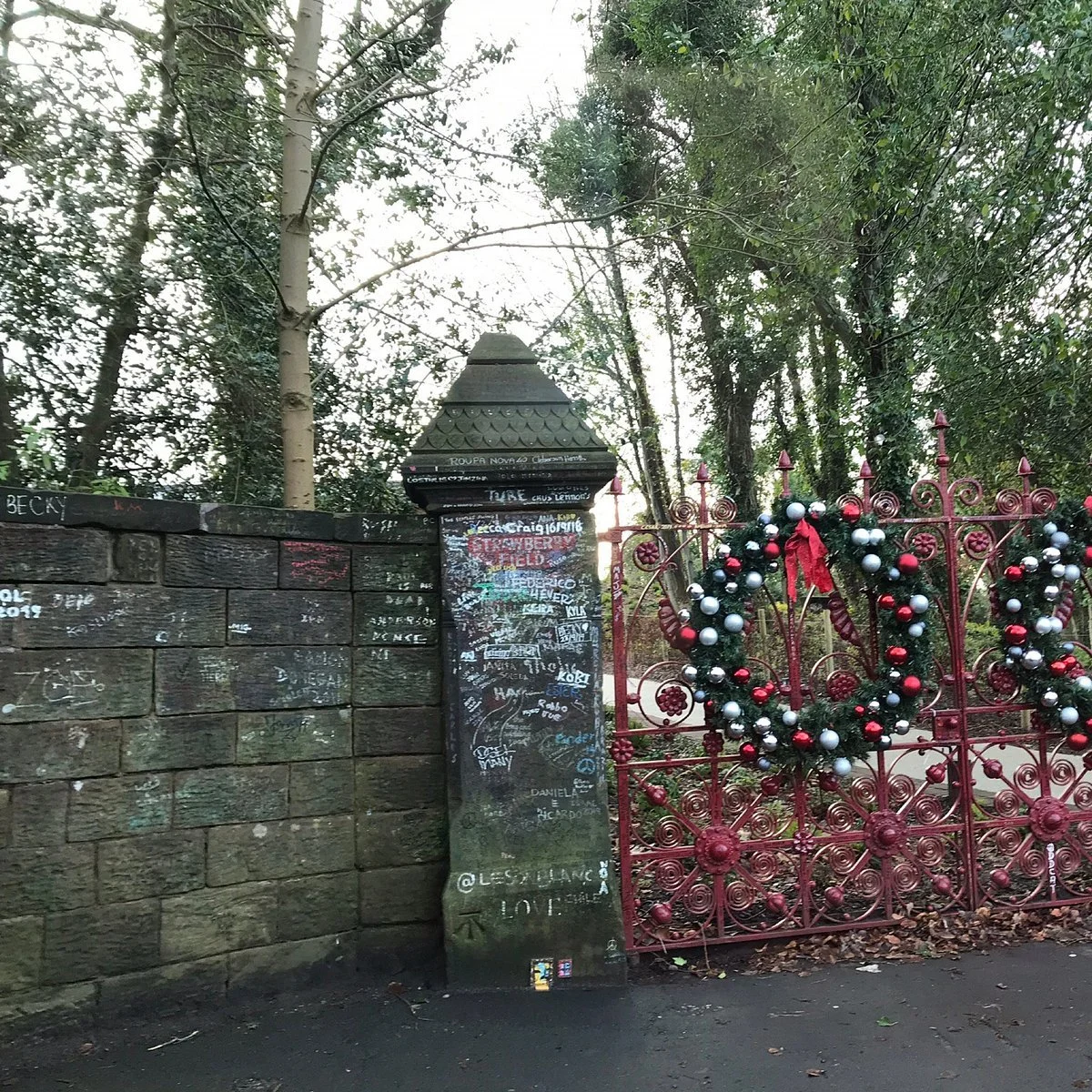 Christmas wreath hanging on the red Strawberry Field gates in Liverpool, with trees and a chalk‑covered stone wall behind