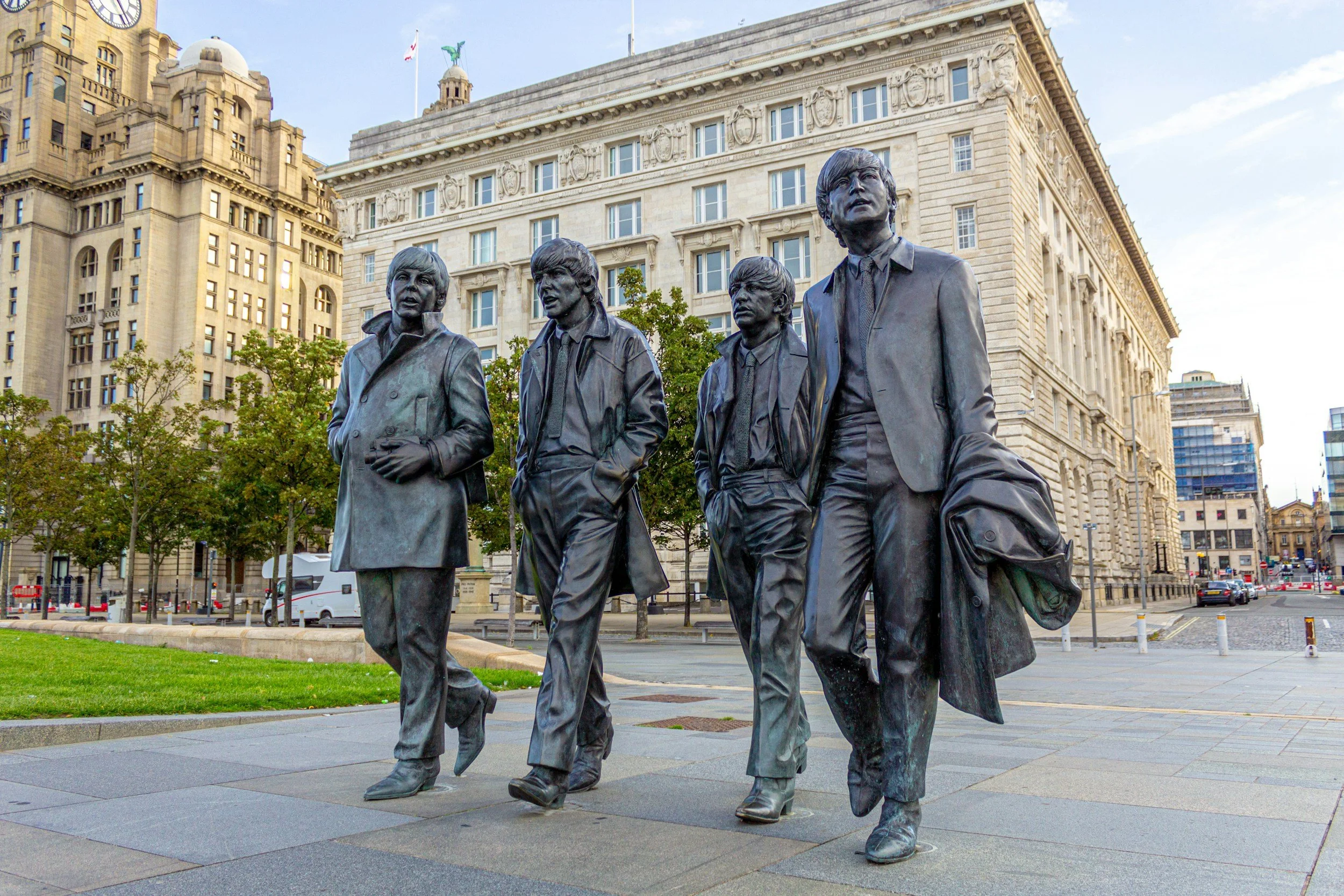Bronze statues of the Beatles, John Lennon, Paul McCartney, George Harrison, and Ringo Starr, standing on a city sidewalk with trees and historic buildings in the background.