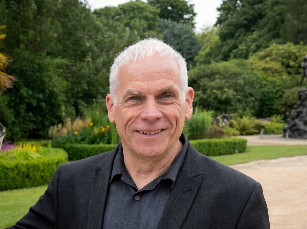 A middle-aged man with short white hair, smiling, standing outdoors in a garden with green trees and bushes in the background.