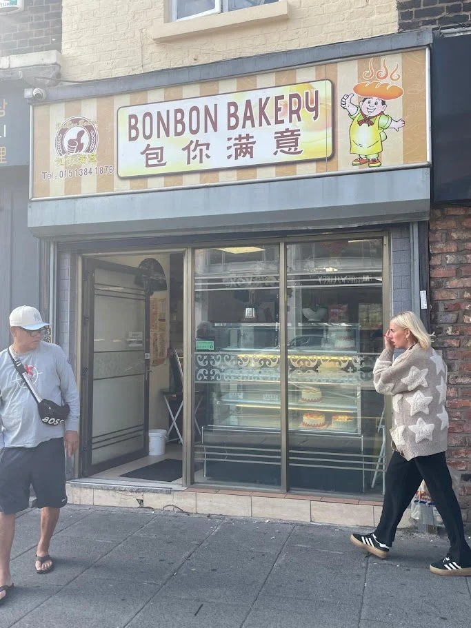 Bonbon Bakery storefront in Liverpool with Chinese‑character signage and a cartoon chef logo, as two people walk past