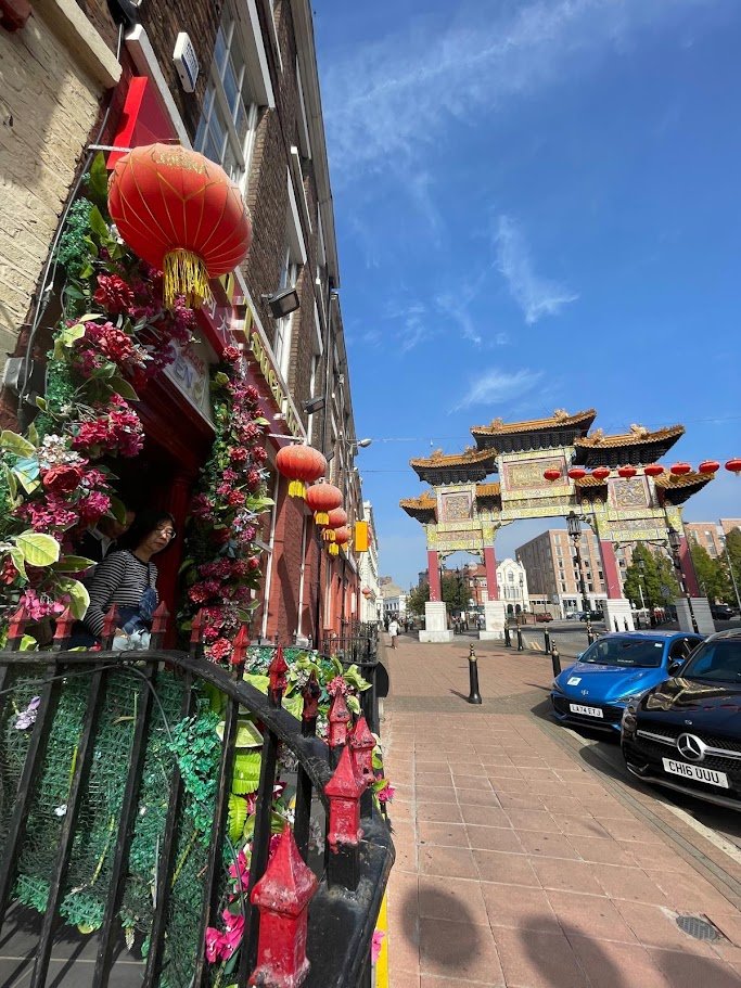 A street scene with a building decorated with red lanterns and colorful flowers, and a traditional Chinese gate in the background under a clear blue sky.
