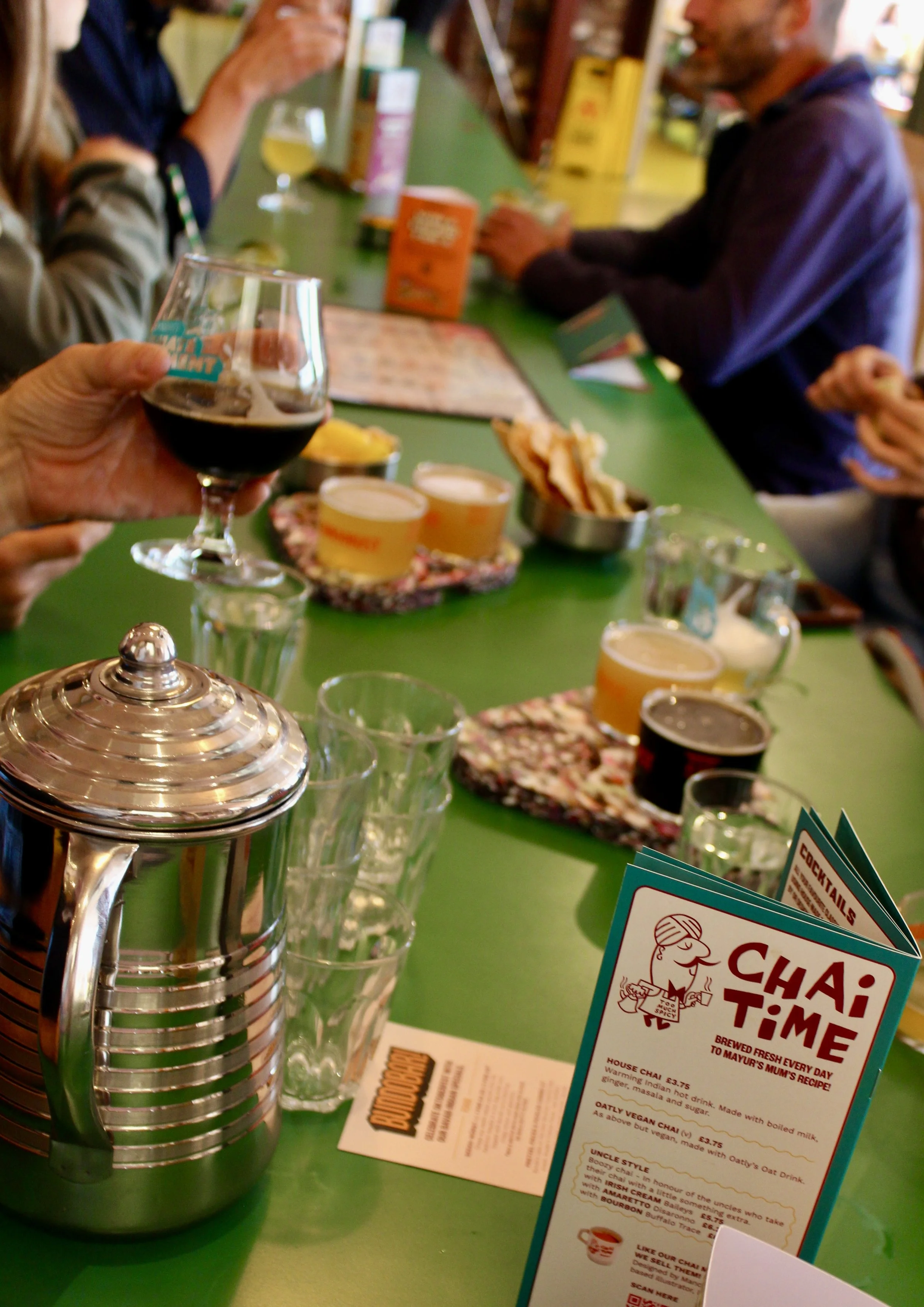 Bar counter in Liverpool with a person holding a glass of dark beer, a shiny metal teapot in the foreground, small bowls of food, stacked glasses and a ‘Chai Time’ menu card