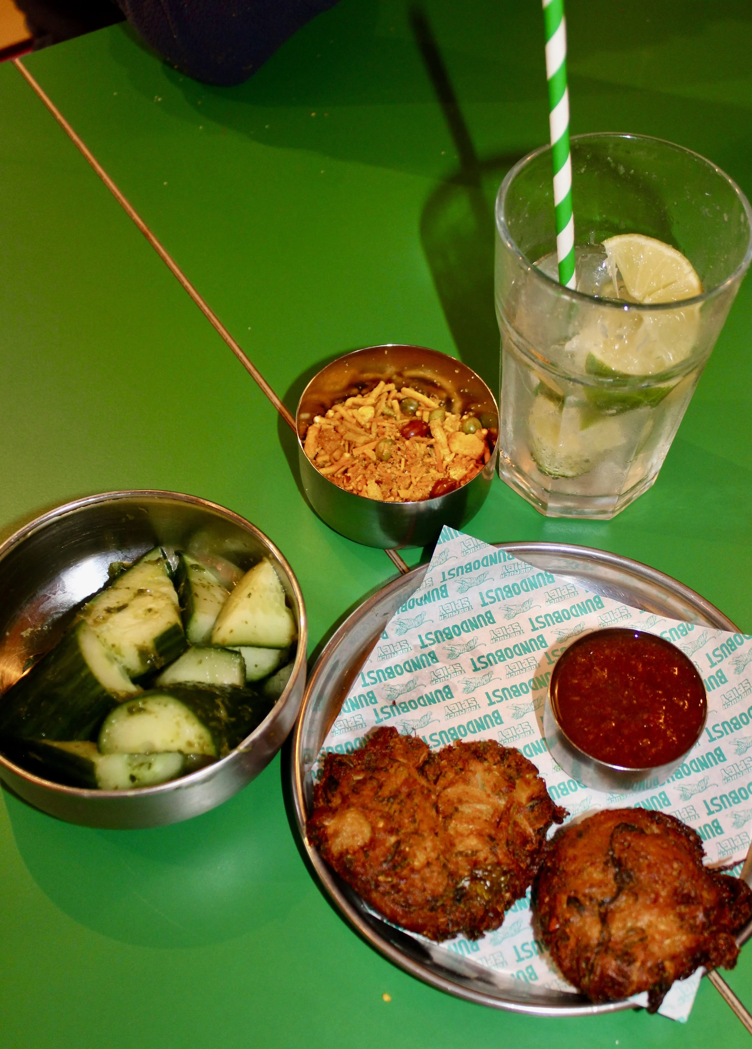 A variety of Indian foods on a green table at Liverpool restaurant, including cucumber slices, fried vegetable cutlets, a glass of lemon water with ice and a striped straw, and a small bowl of spicy red chutney.