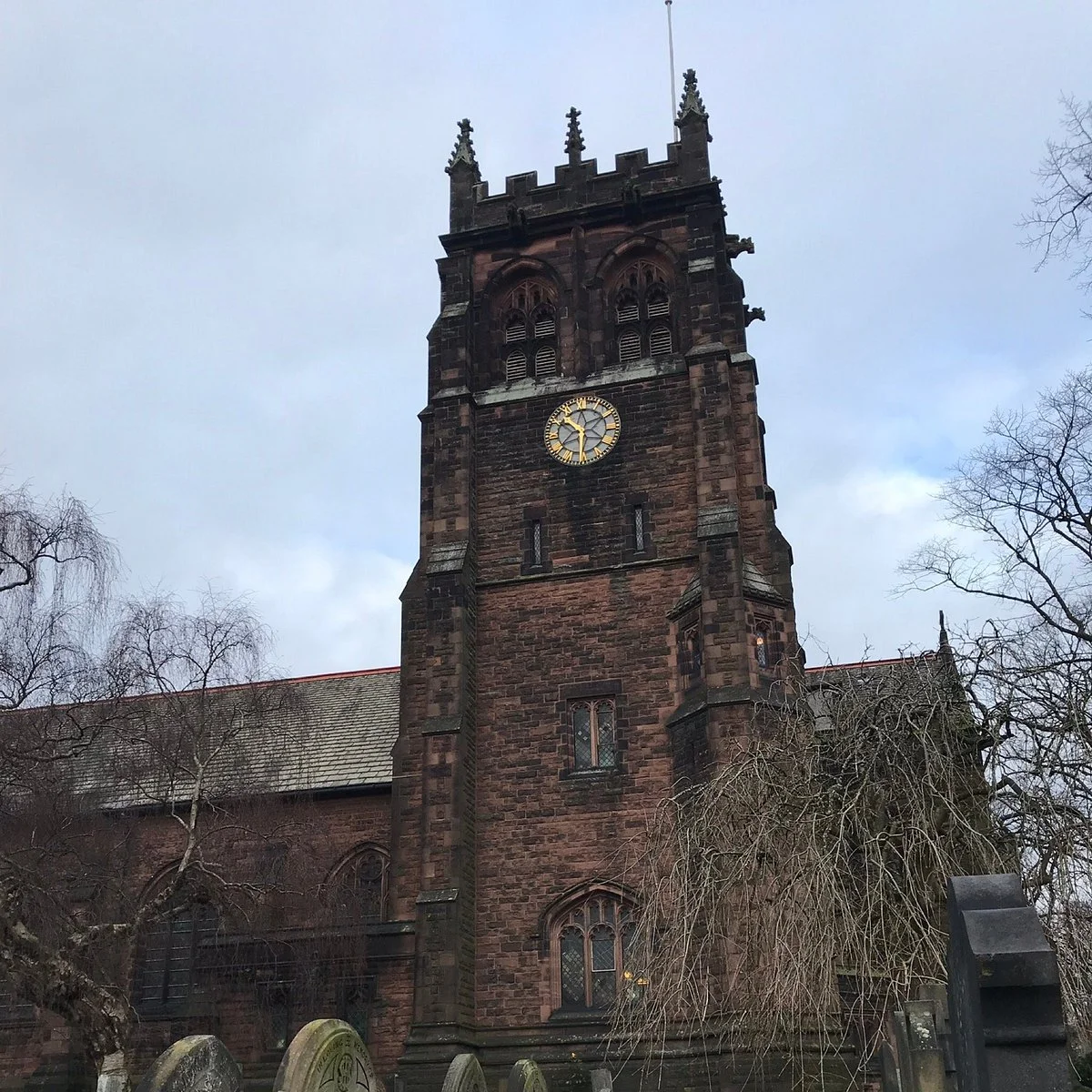 Historic stone church tower in Woolton, Liverpool, surrounded by leafless trees under a cloudy sky