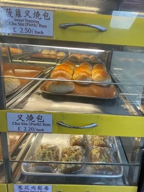 Display case with baked goods including sweet topping cha siu pork buns on the upper shelf and assorted buns on the lower shelf in a Liverpool bakery
