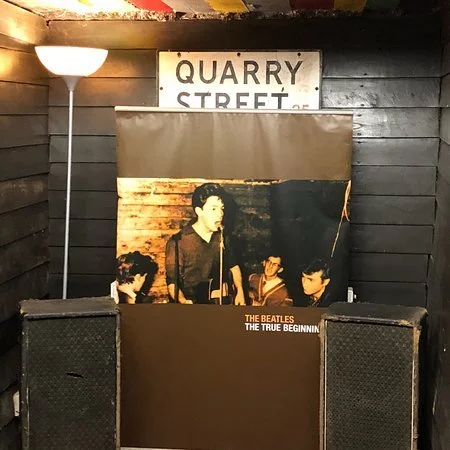Dimly lit room with a vintage Beatles poster, two large speakers, a Quarry Street sign and a floor lamp against a brick wall.