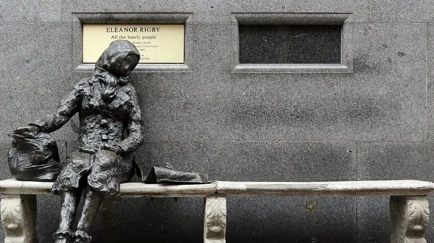Bronze statue of Eleanor Rigby sitting on a bench with her head resting on her hand beside a bag, in front of the Eleanor Rigby plaque in Liverpool