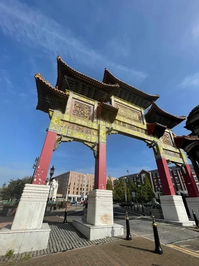 A traditional Chinese gate with ornate designs, red pillars, and a multi-tiered roof, set against a blue sky in an urban area.