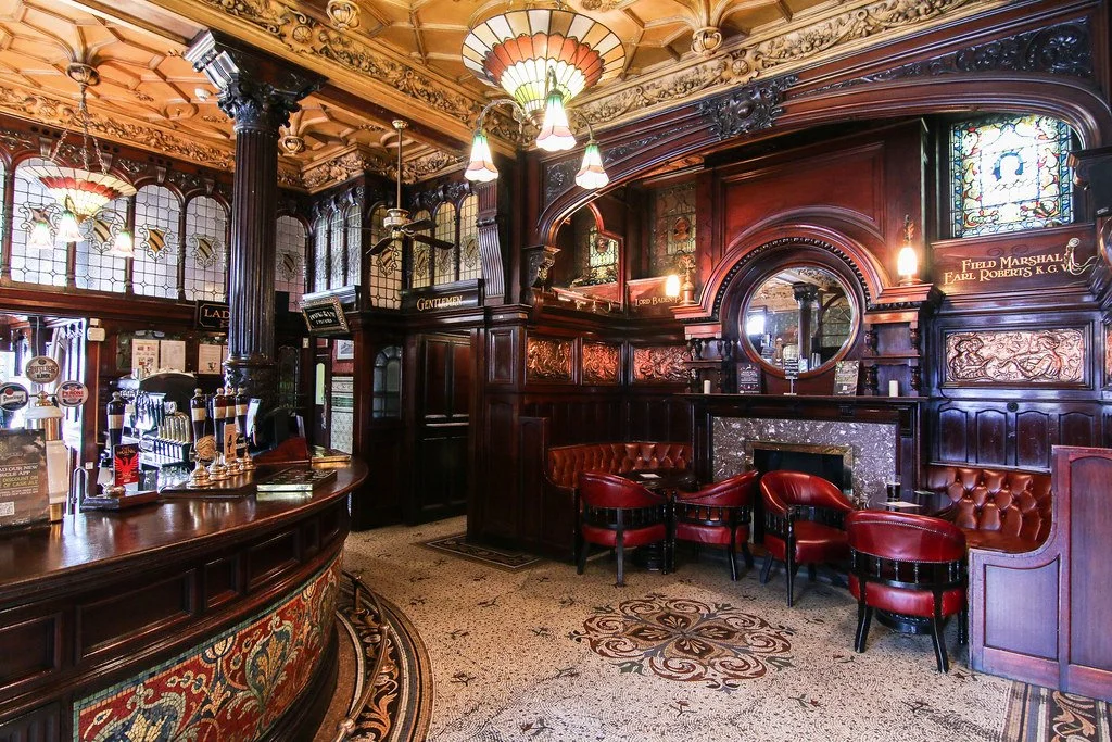 Vintage‑style Liverpool bar at The Philharmonic Dining Rooms pub interior with dark wood panelling, stained‑glass windows, red leather chairs and ornate decorative details