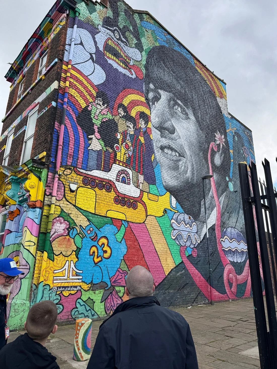 Colourful mural on a Liverpool brick building featuring a black‑and‑white portrait of Ringo Star surrounded by cartoon characters and abstract patterns