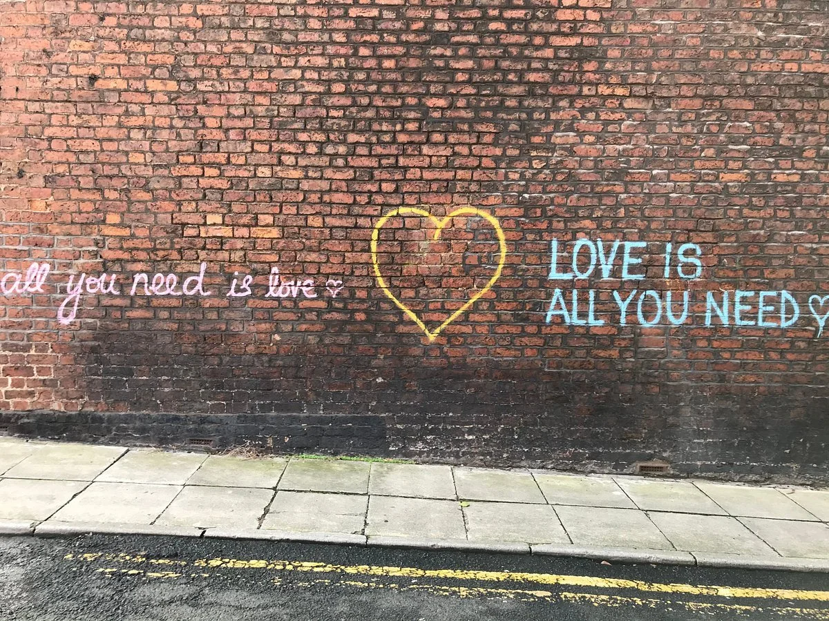 Beatles‑themed graffiti on a Liverpool brick wall with a yellow heart, pink ‘all you need is love’ text and blue ‘LOVE IS ALL YOU NEED’ lettering