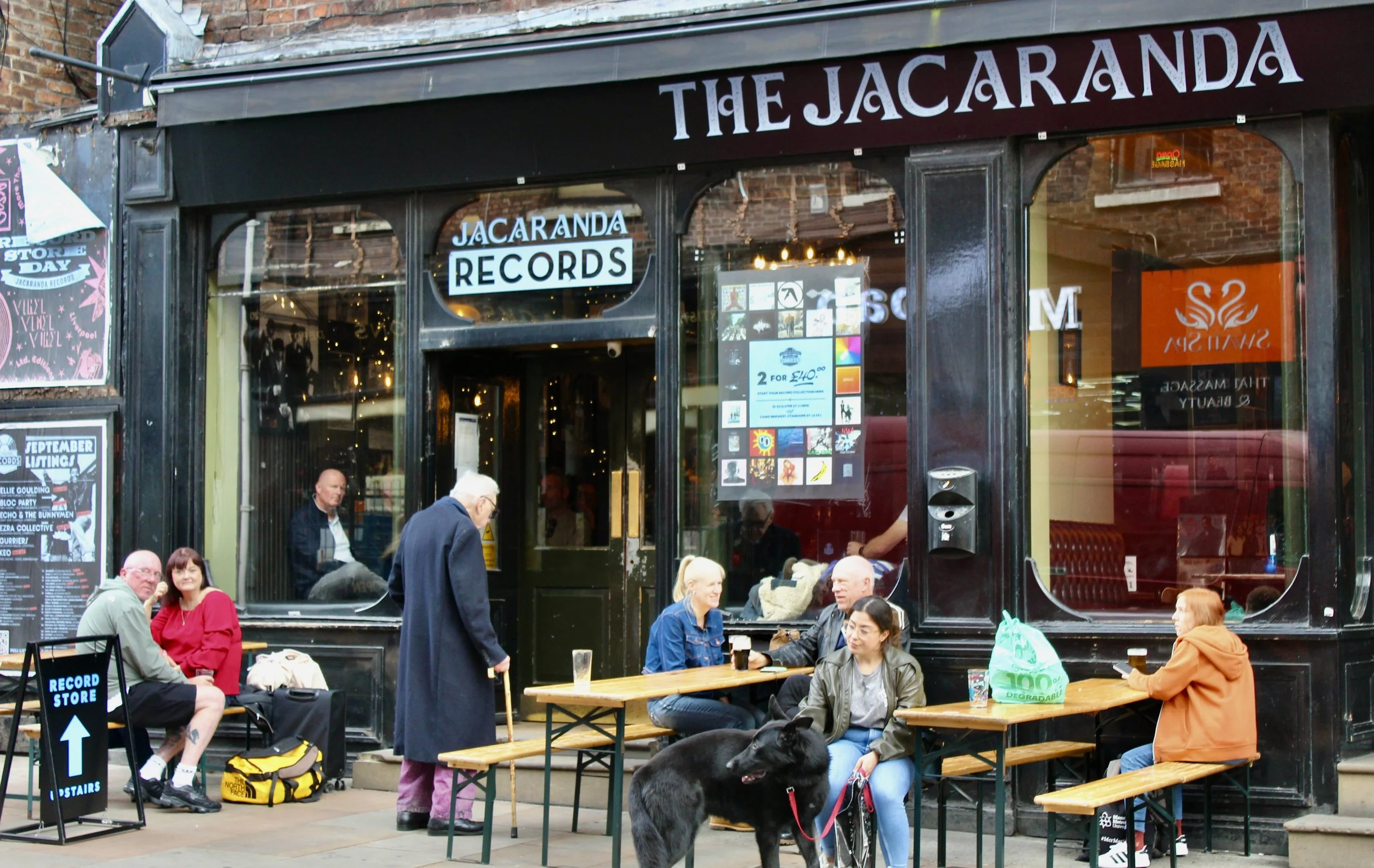 People sitting and standing outside The Jacaranda in Liverpool, with tables, benches, posters on the windows and a dog by the entrance