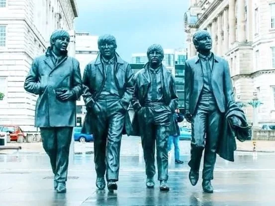 Statue of the four Beatles walking together in a city street with old buildings and cars in the background.
