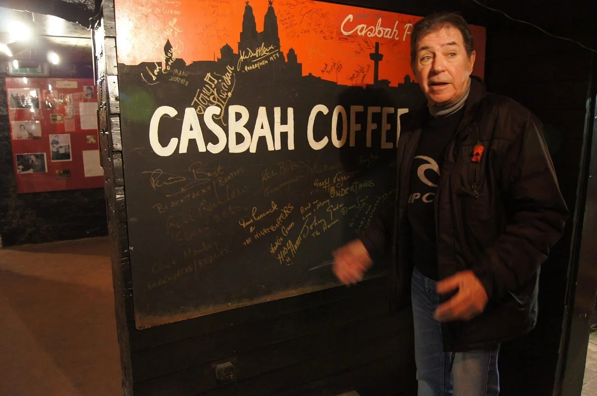 Man standing beside the Casbah Coffee Club sign covered in signatures, with a cathedral silhouette and cross in the background in Liverpool
