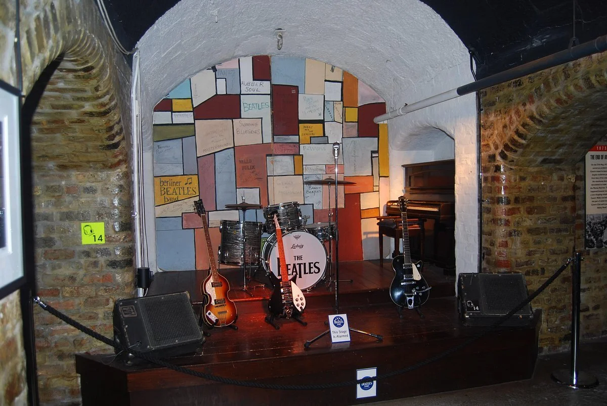 Music stage in a Liverpool venue with drums, electric guitars, a piano and a microphone, set against a colourful abstract mural with ropes marking restricted access where the Beatles played.