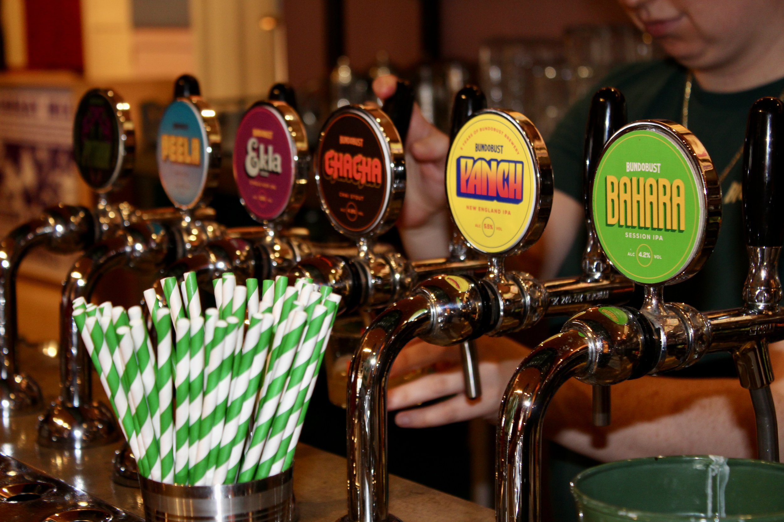 Close‑up of colourful beer tap handles at a Liverpool bar, with a person in the background and a container of green‑and‑white striped straws in the foreground