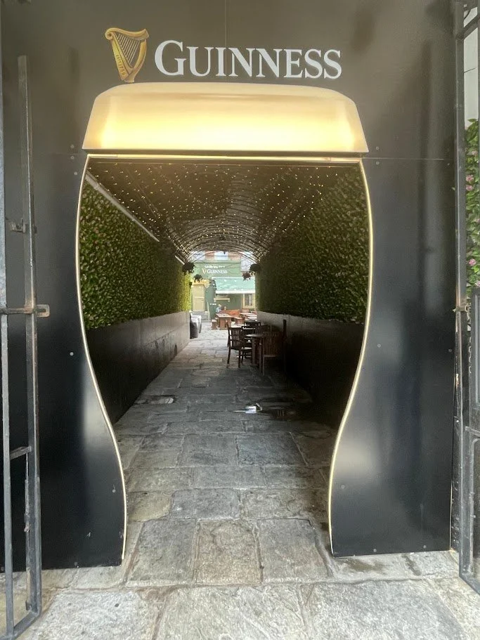 Guinness‑glass‑shaped entrance arch leading to a Liverpool outdoor patio with tables, chairs, greenery and overhead lights