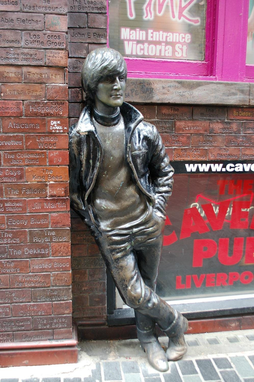 Bronze statue of Billy Fury in a leather jacket leaning against a brick wall outside a Liverpool music venue with pink and red signage