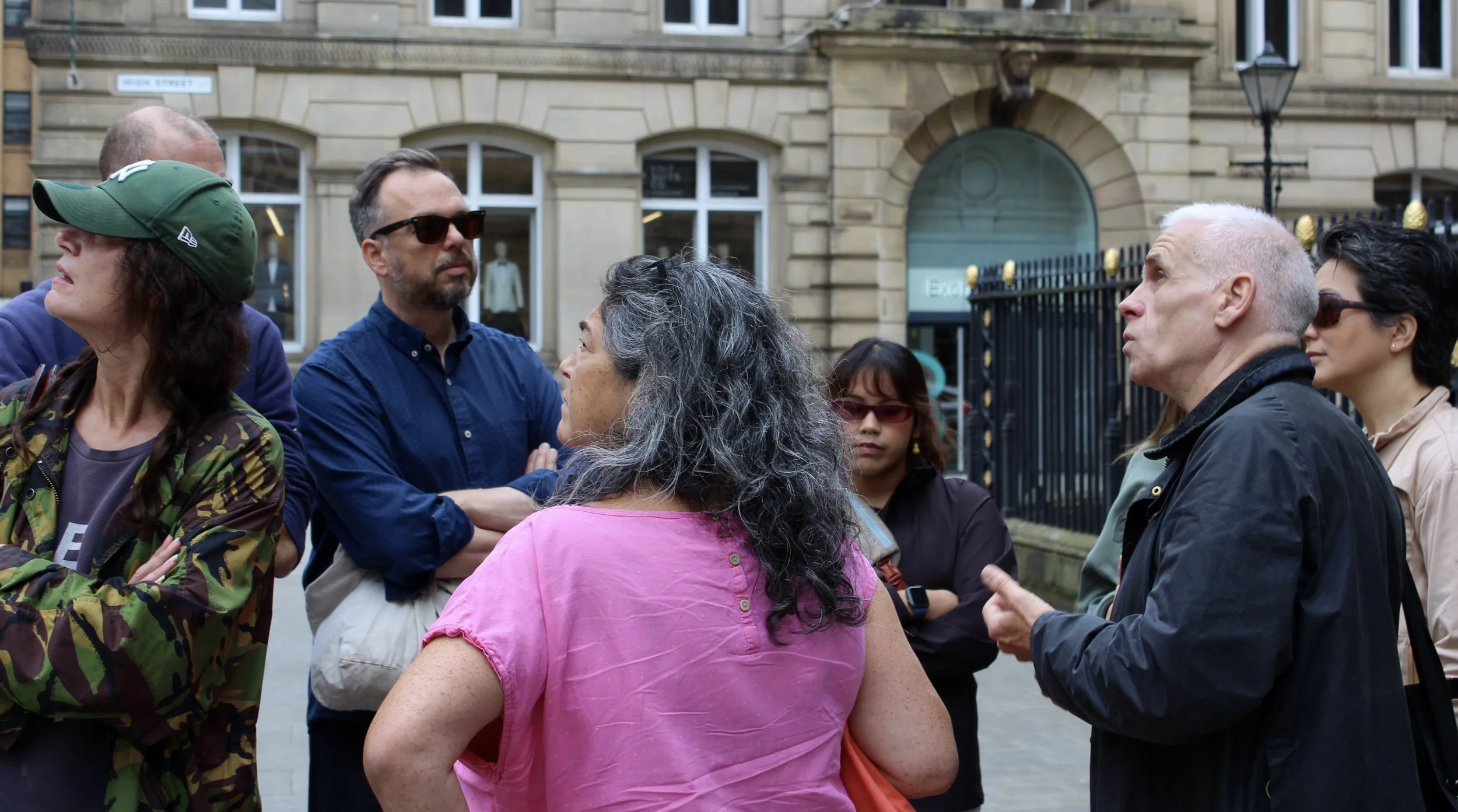 Group of diverse people engaged in conversation outside in an urban setting with a historical building and black iron fence in the background.