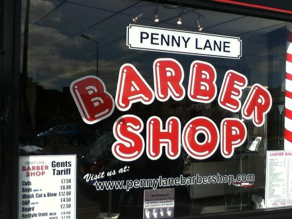 Storefront window of the Penny Lane Barber Shop in Liverpool with red and white lettering and a gents grooming price sign