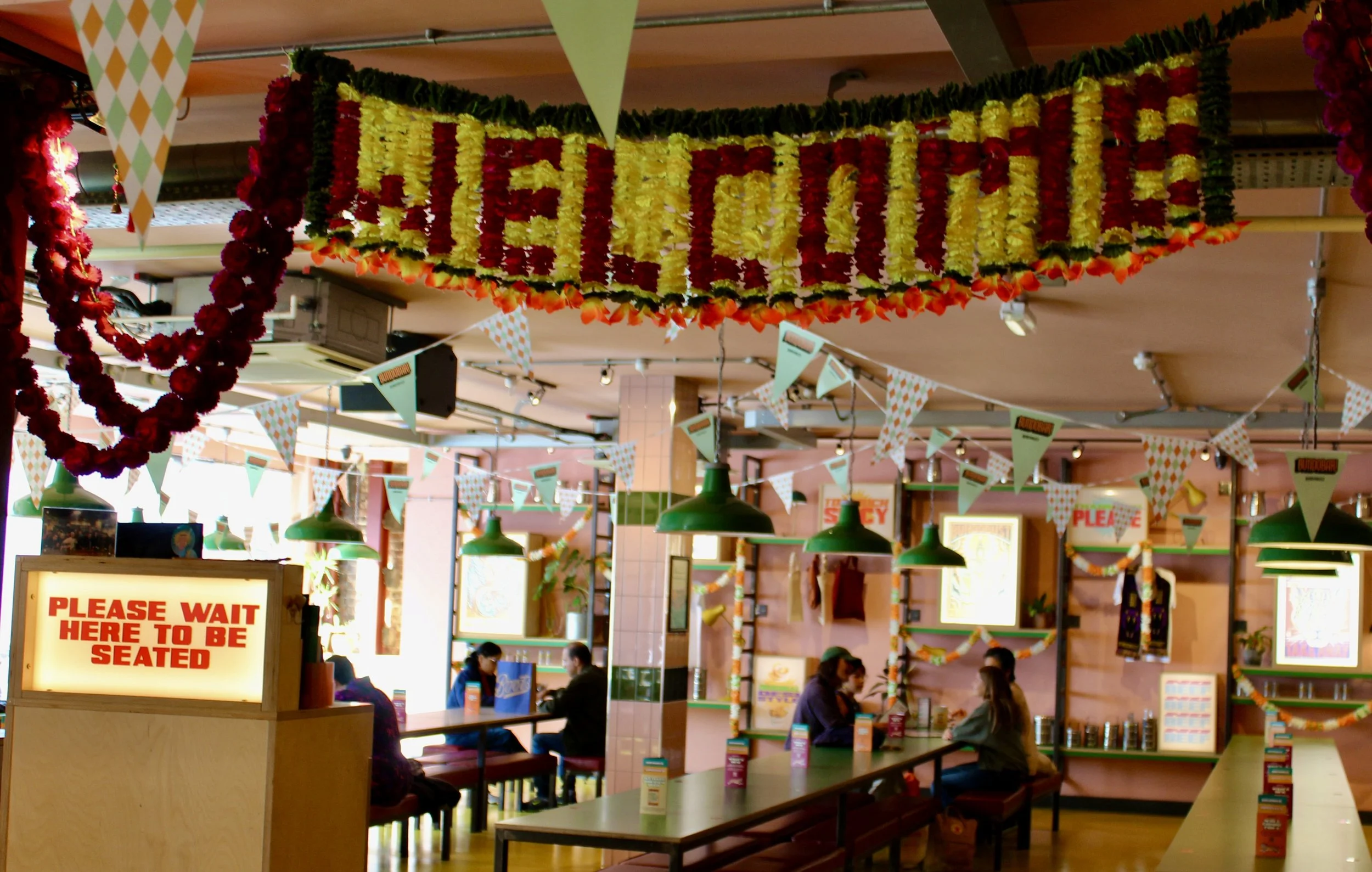 Indoor Liverpool dining space decorated with yellow and red flower arrangements, bunting and hanging signs, with people sitting at tables and waiting in line
