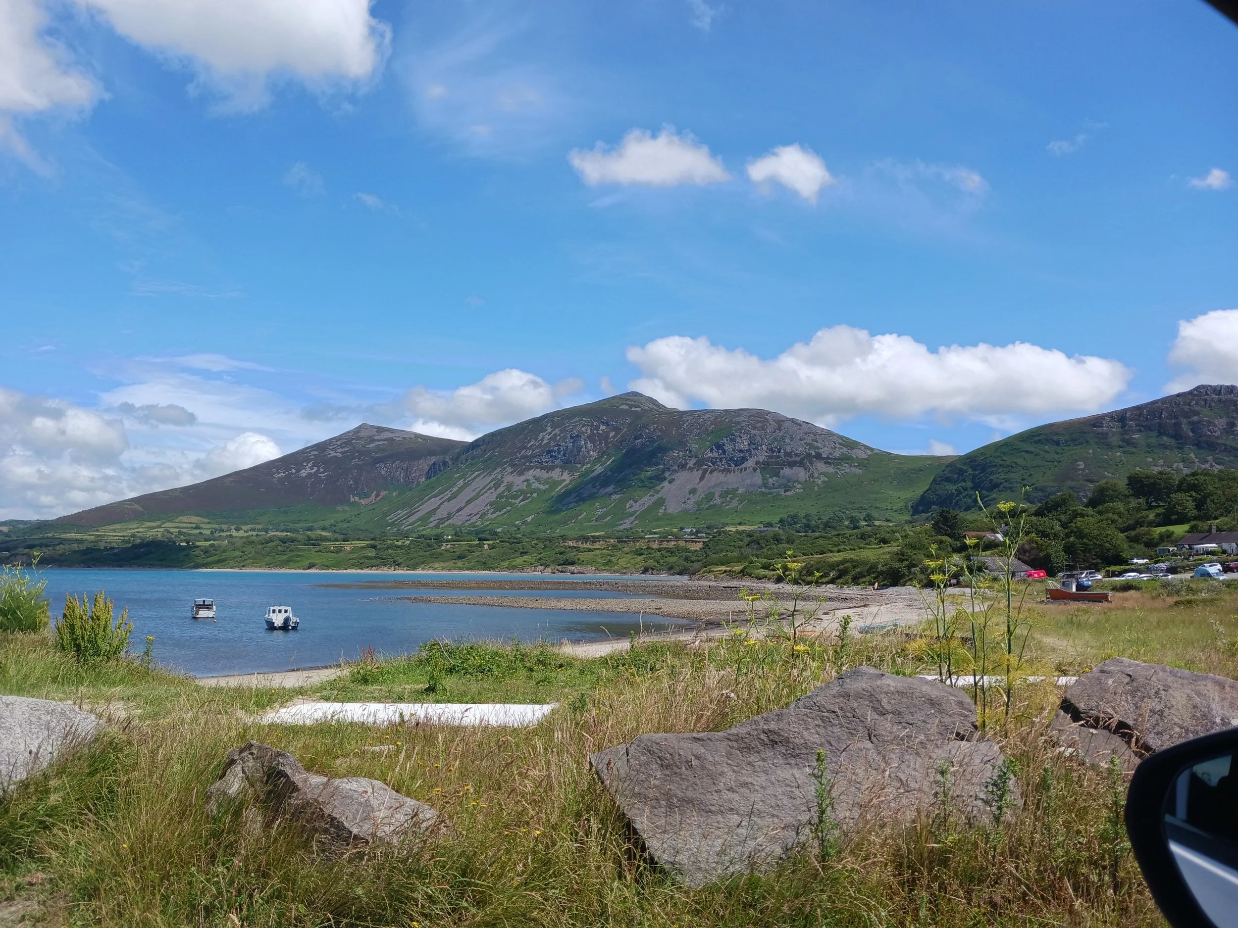 Scenic view of a lake with boats, surrounded by green hills and mountains, under a blue sky with white clouds.