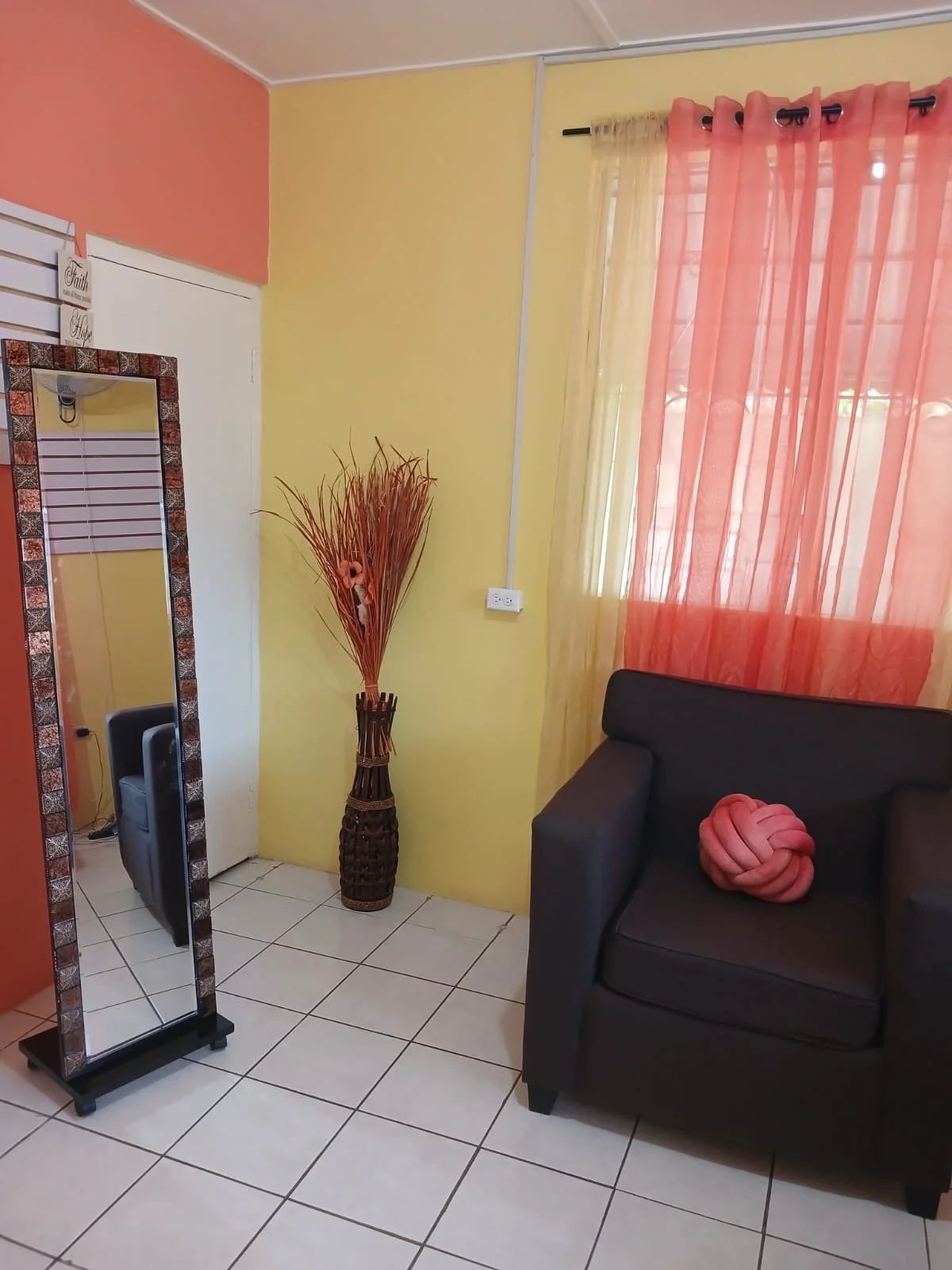 Interior of a cozy living room with a black sofa, pink decorative pillow, yellow and peach-colored walls, sheer pink and yellow curtains, a tall wicker vase with dried branches, a full-length mirror with a decorative frame, and white tiled floor.