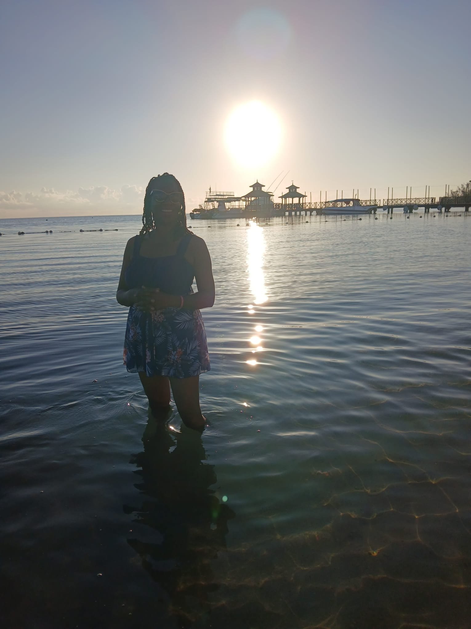 A woman stands in shallow water at sunset with an ocean pier and boats in the background.