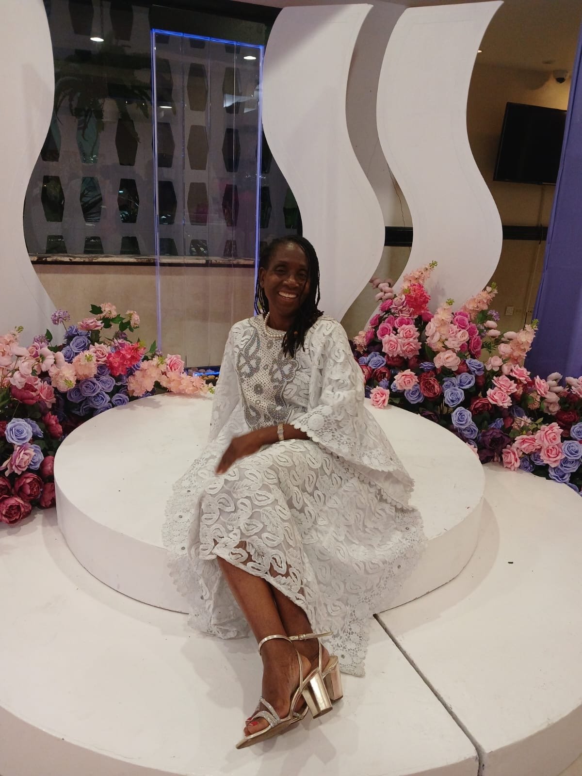 Woman in a white lace dress sitting on a round white platform, surrounded by pink and purple flowers, in an elegant indoor setting.