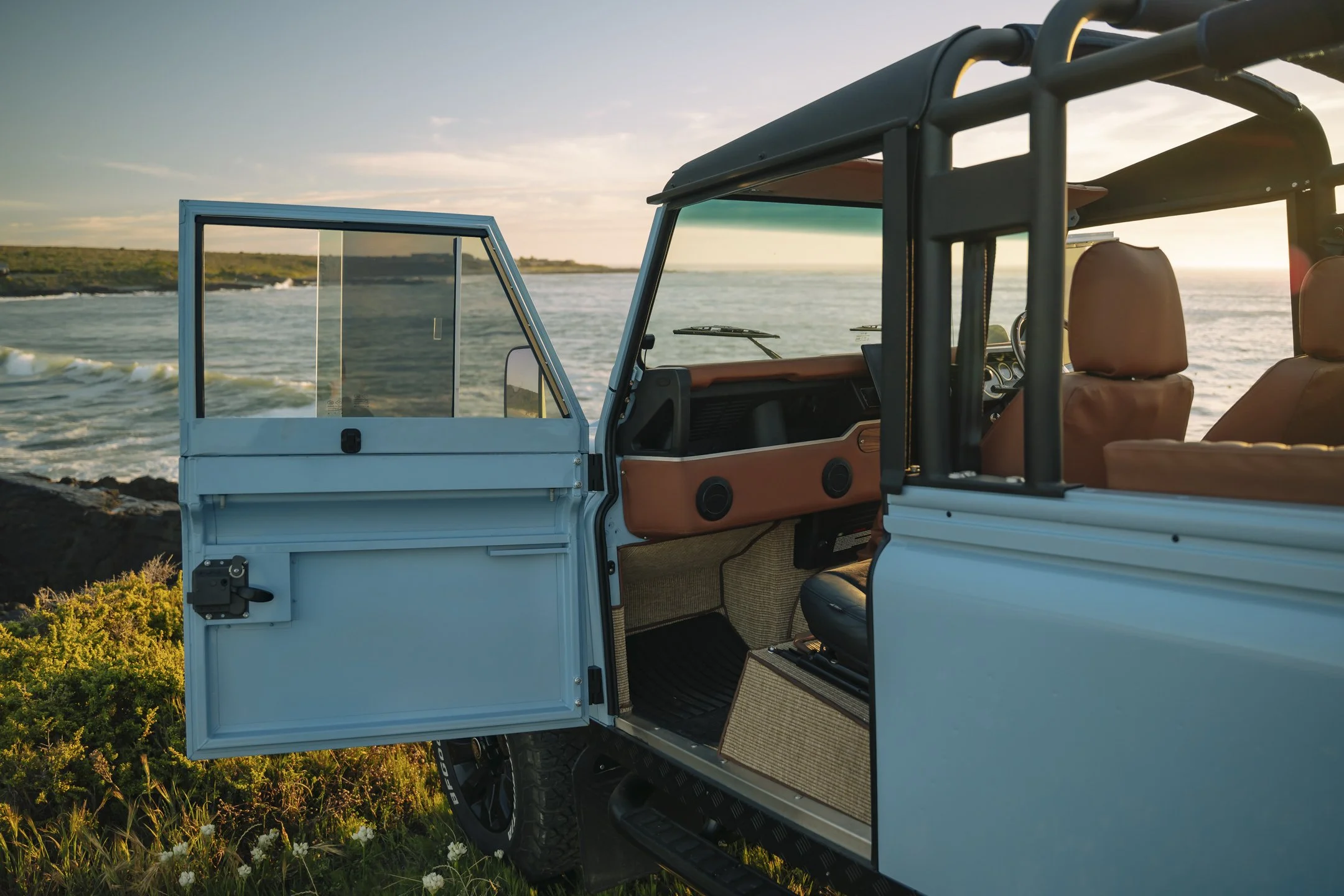 A partially opened light blue off-road vehicle parked on grassy terrain near water, with coastline and sky in the background during sunset.