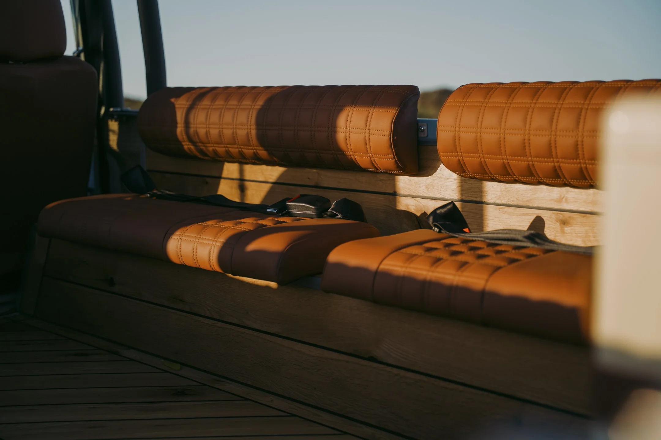 Inside a vehicle with tan quilted leather seats and a wooden interior, showing sunlight streaming in.