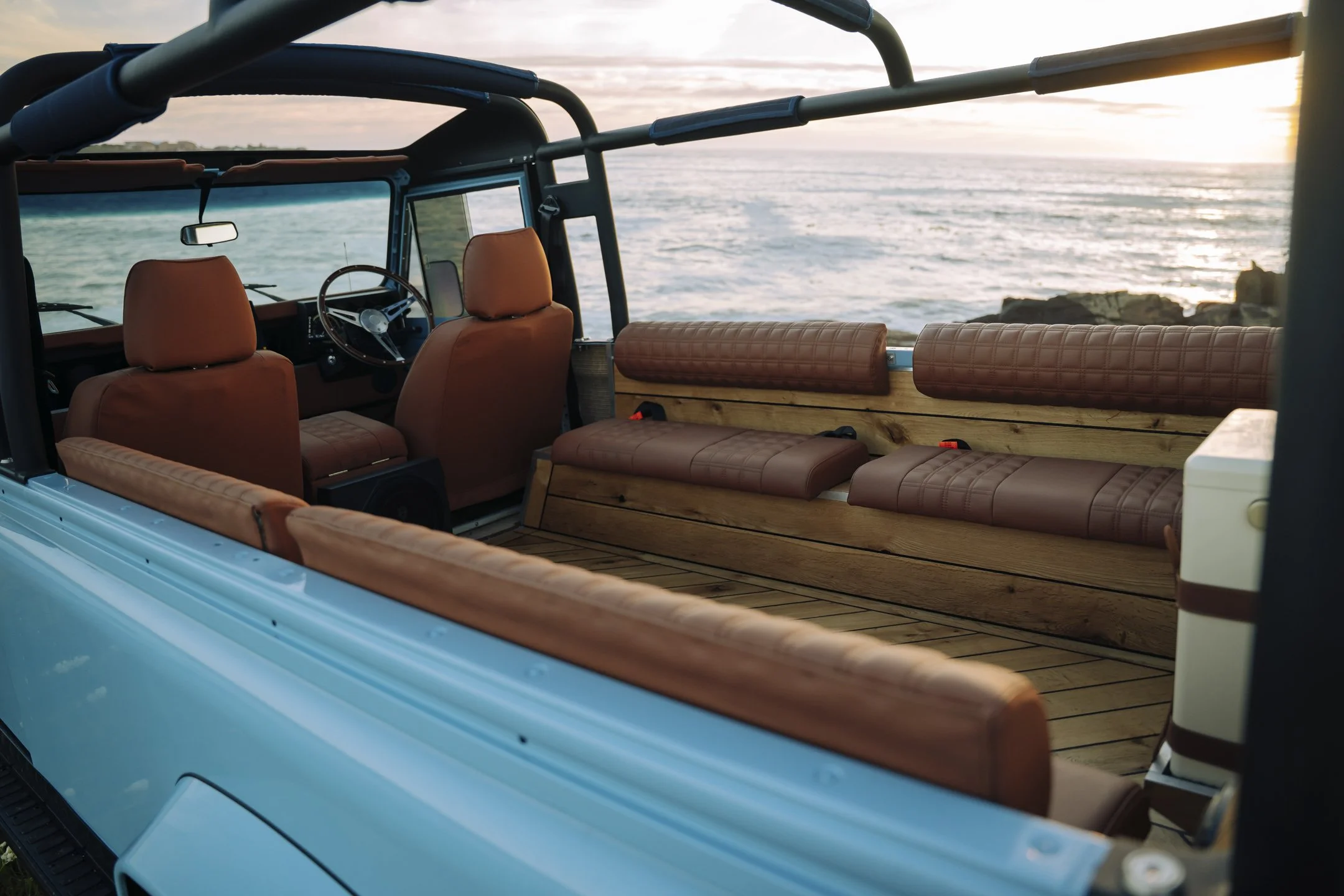 Interior of a vintage off-road vehicle with brown leather seats and a wooden panel, overlooking the ocean during sunset.